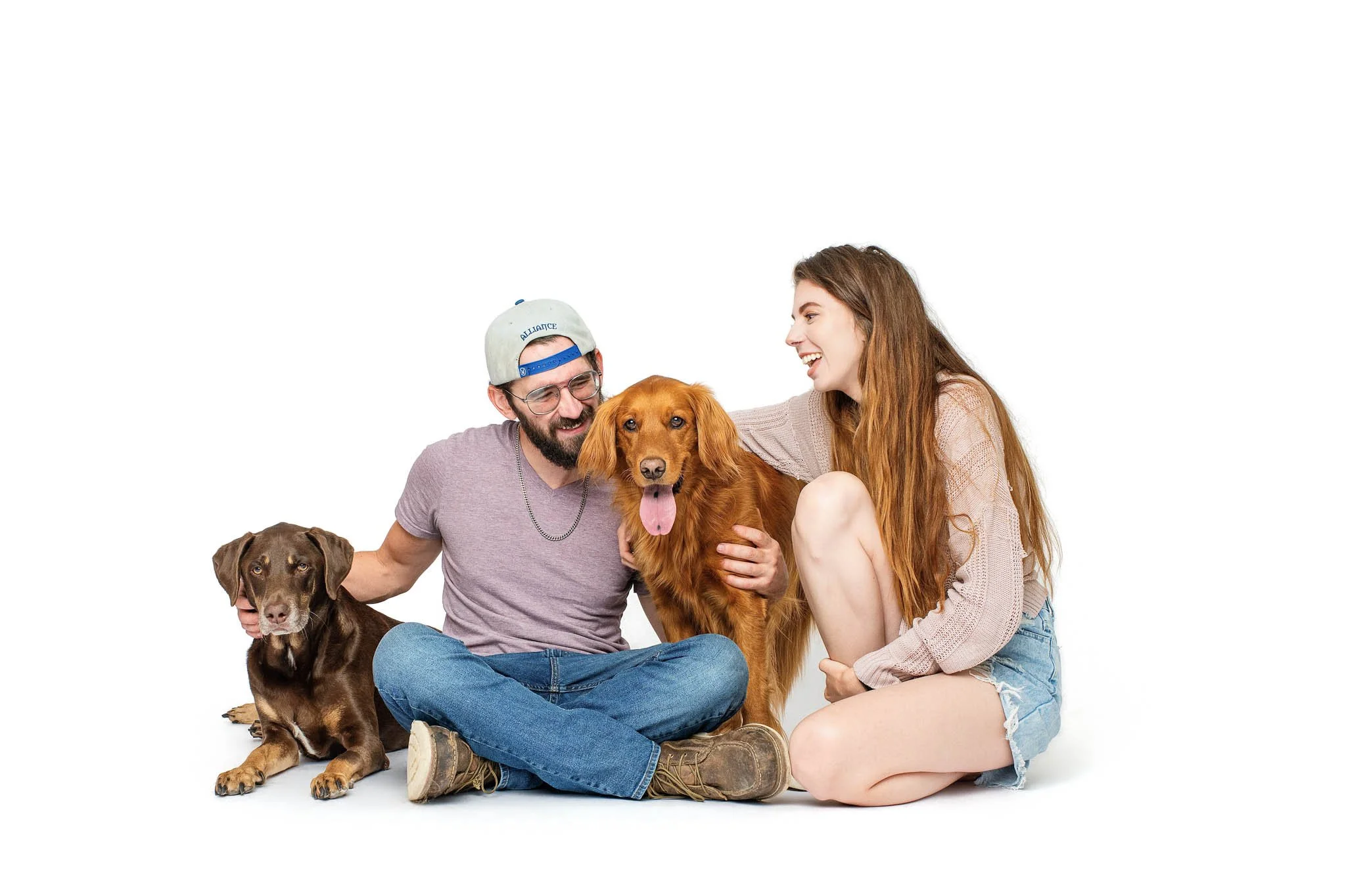 A smiling couple sits on a white studio floor with Lilah, a long coated red dog, between them and Hank, a chocolate German Shorthaired Pointer and Labrador mix, lying at the man's side.