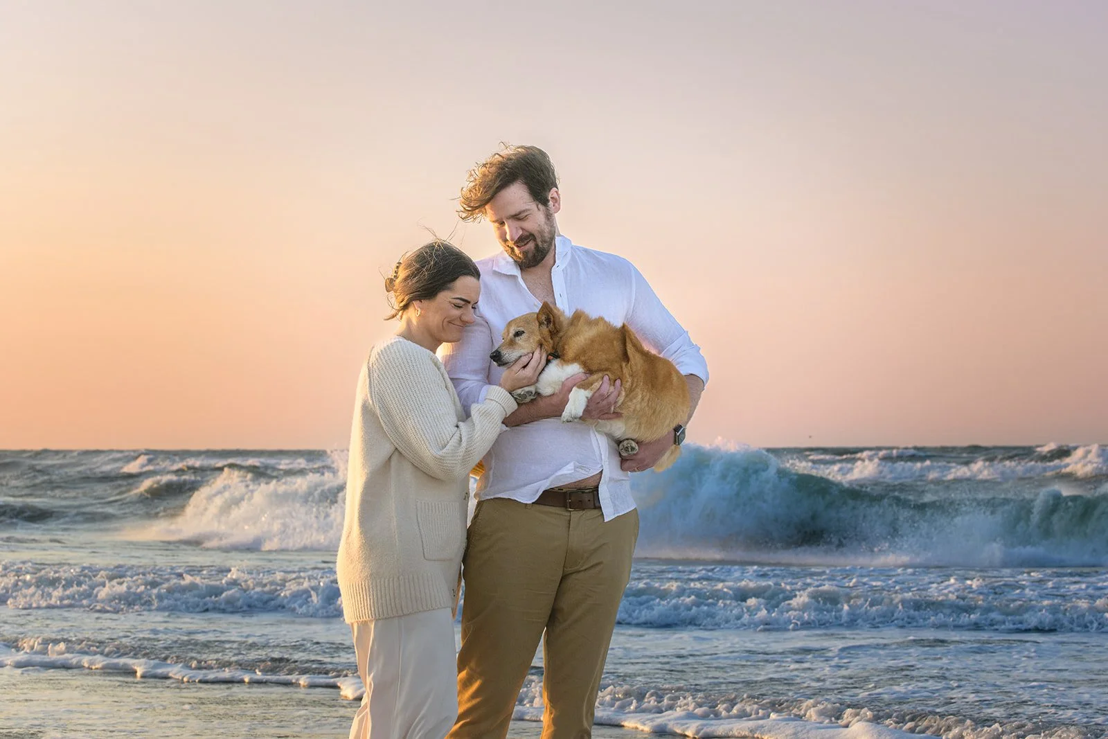 Couple holding a dog on a beach at sunset with ocean waves behind them