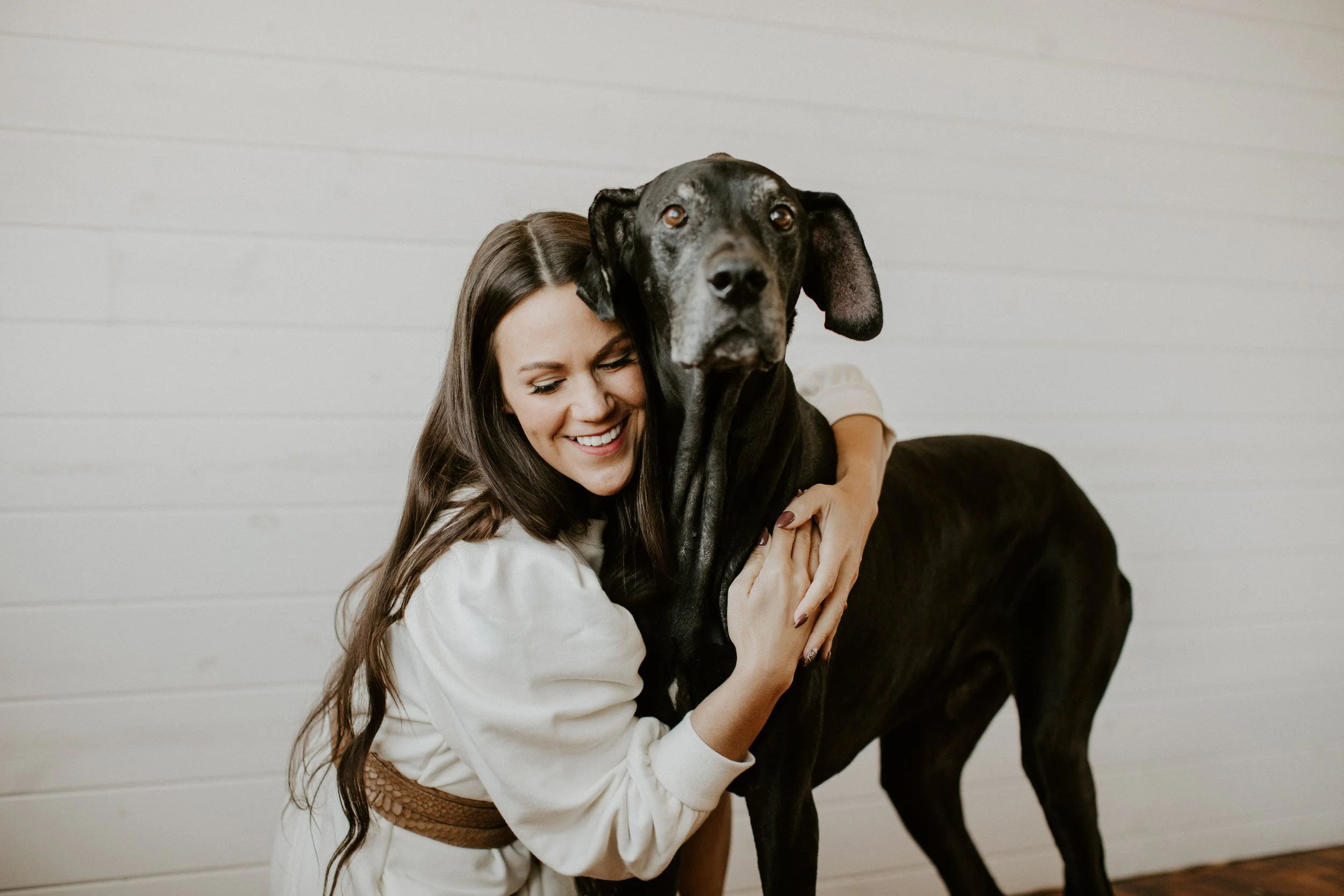 Smiling woman hugging a large black dog indoors against a white wall