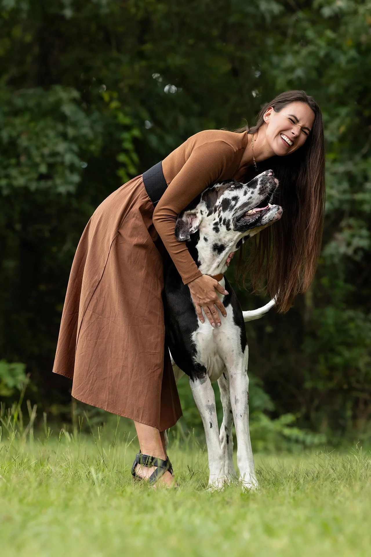 Smiling woman hugging a happy black-and-white Great Dane outdoors in a forest clearing