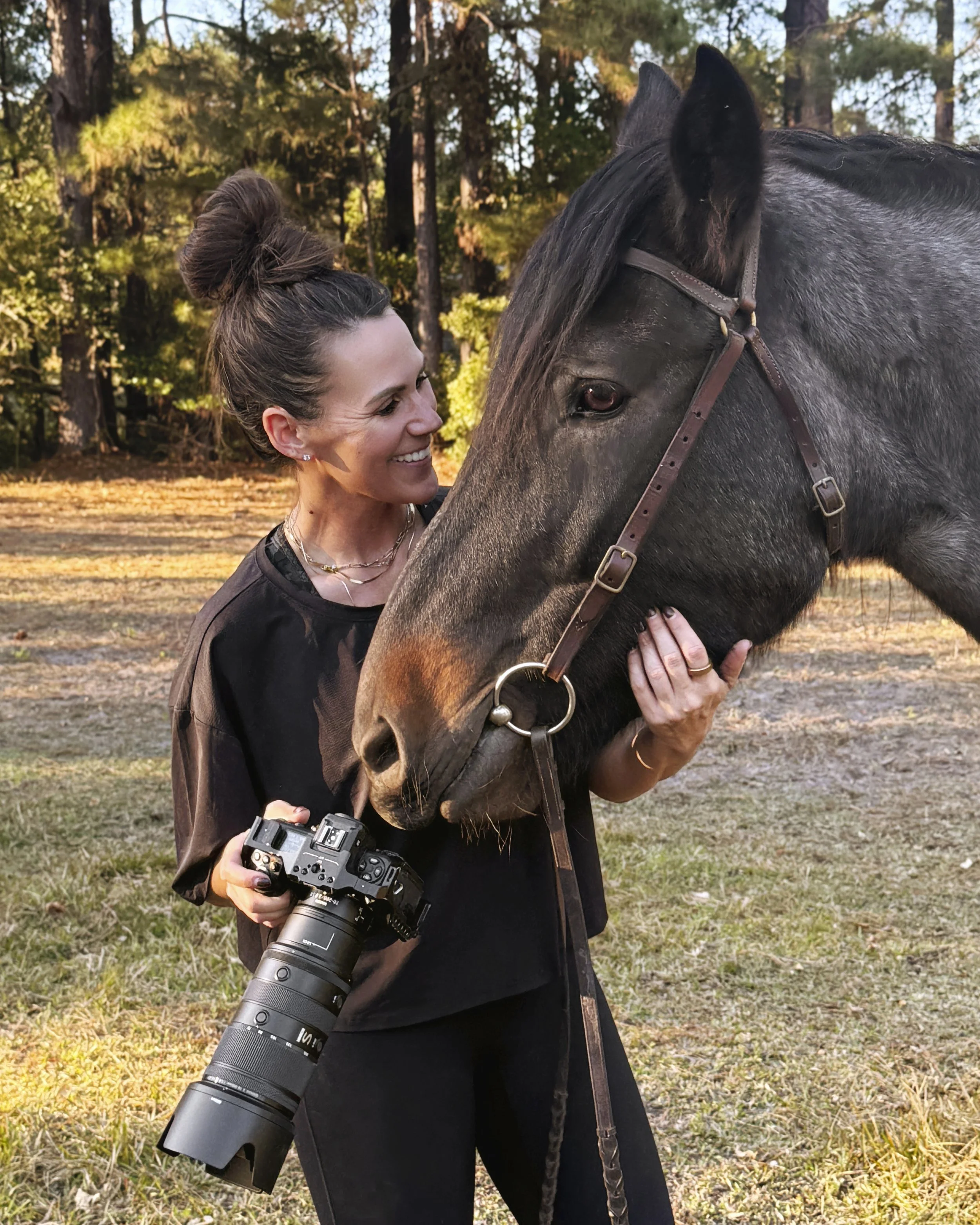 Smiling woman holding a camera and embracing a black horse in a sunlit wooded area