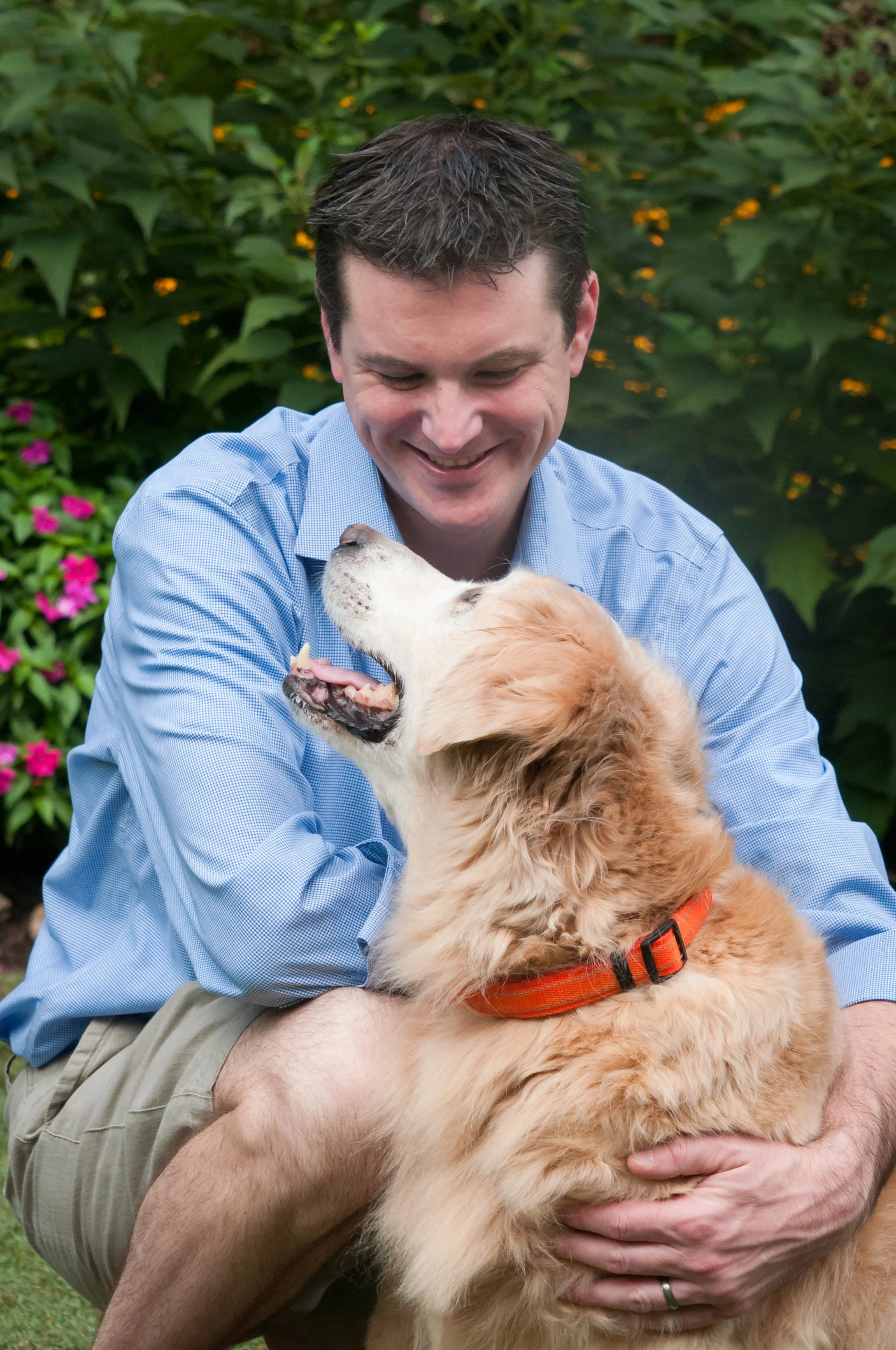 Smiling man in a blue shirt cuddling a fluffy golden dog in a flower garden