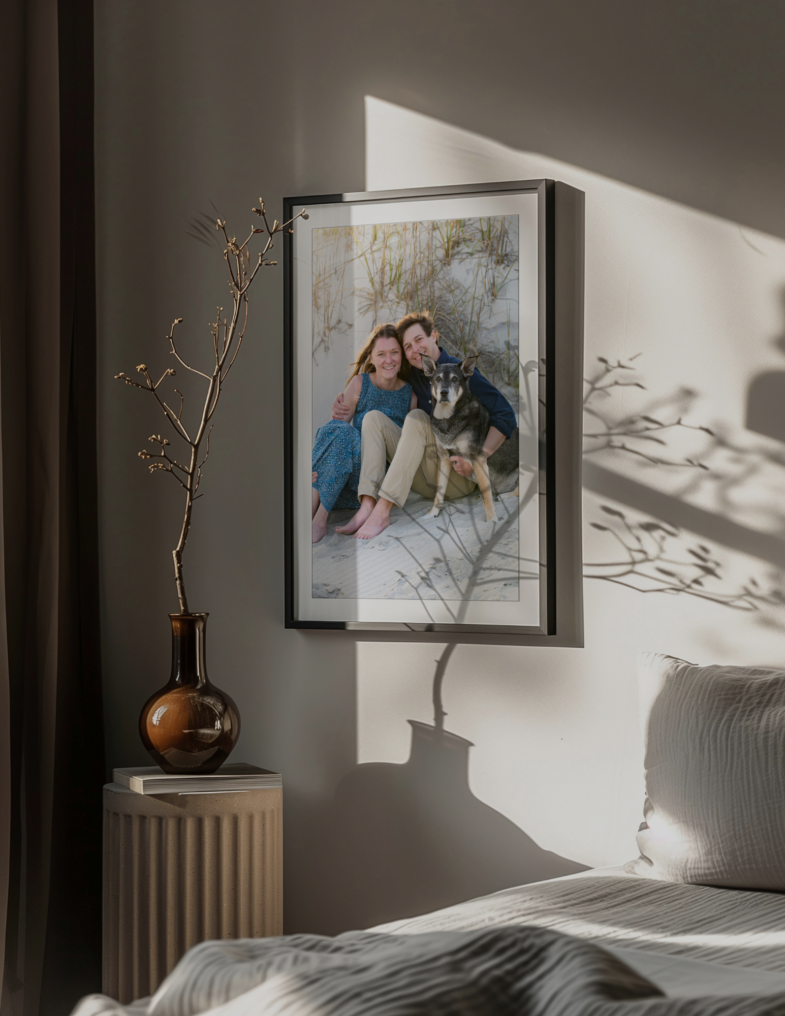 Framed photo of a couple sitting with their dog above a bedroom nightstand with a vase and dried branch
