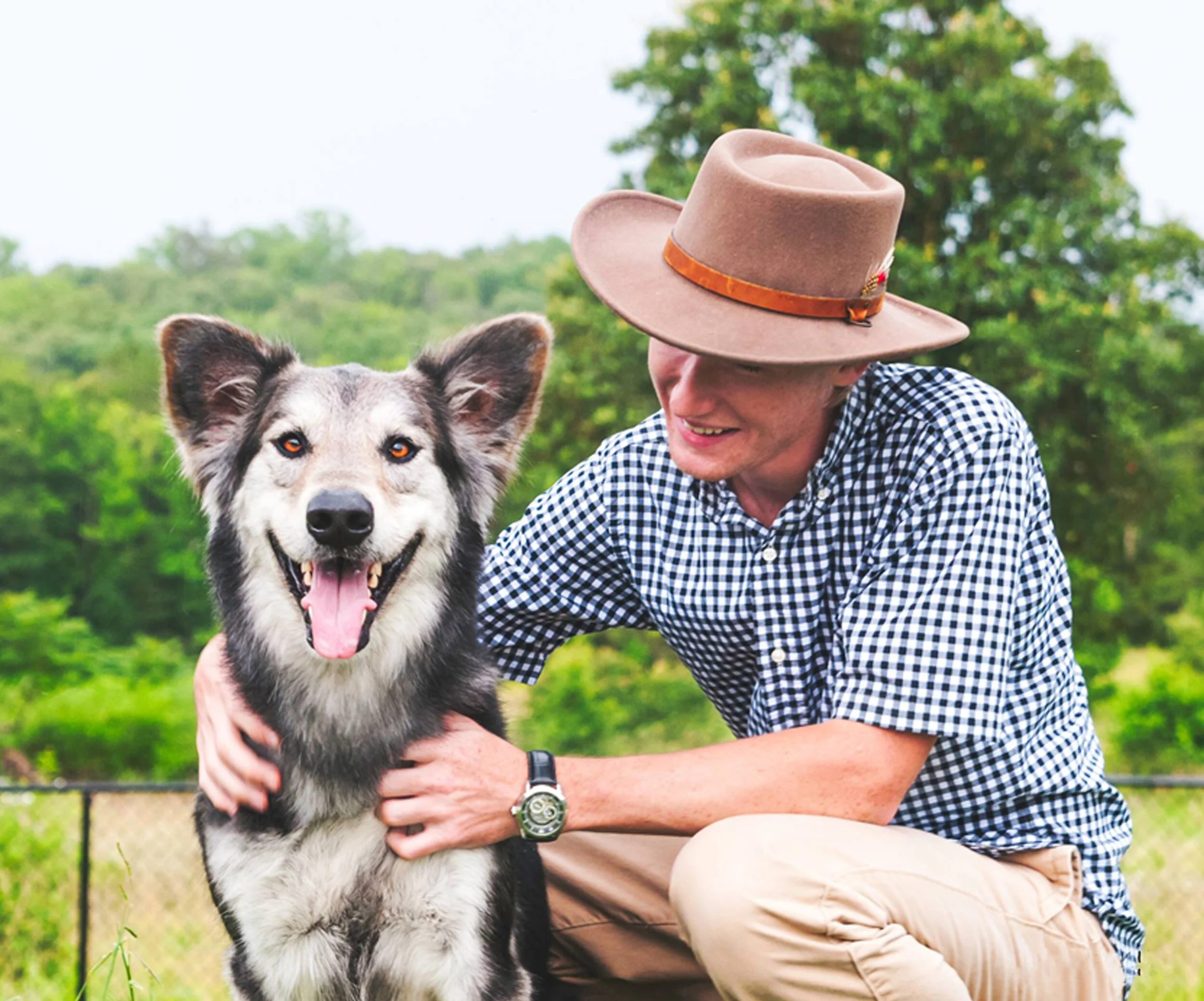 A man in a felt hat and navy gingham shirt kneels beside his smiling black and silver dog in a green field.