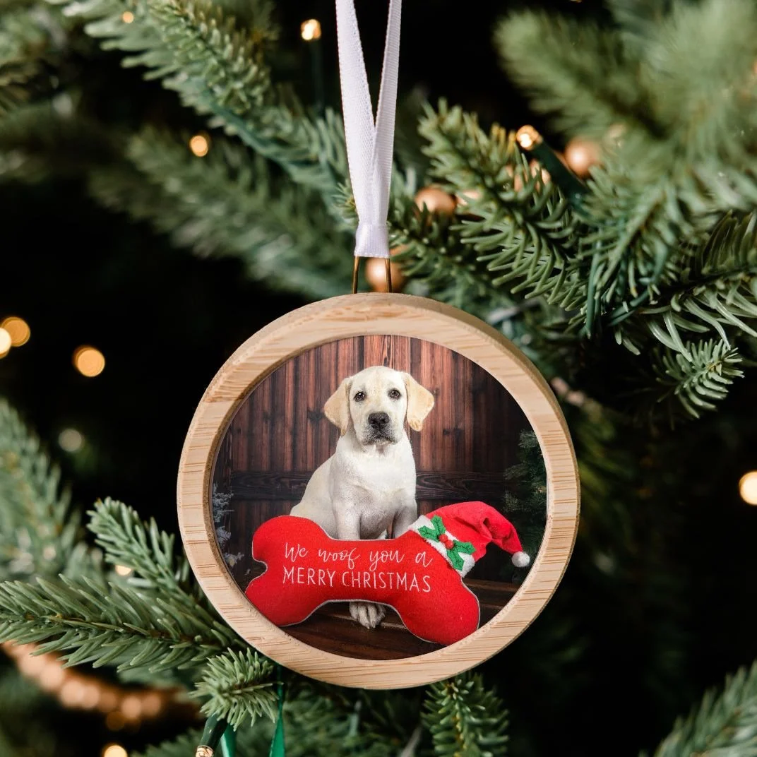 Round bamboo ornament with a portrait of a yellow Lab puppy beside a red bone pillow reading We woof you a Merry Christmas, hanging on a Christmas tree