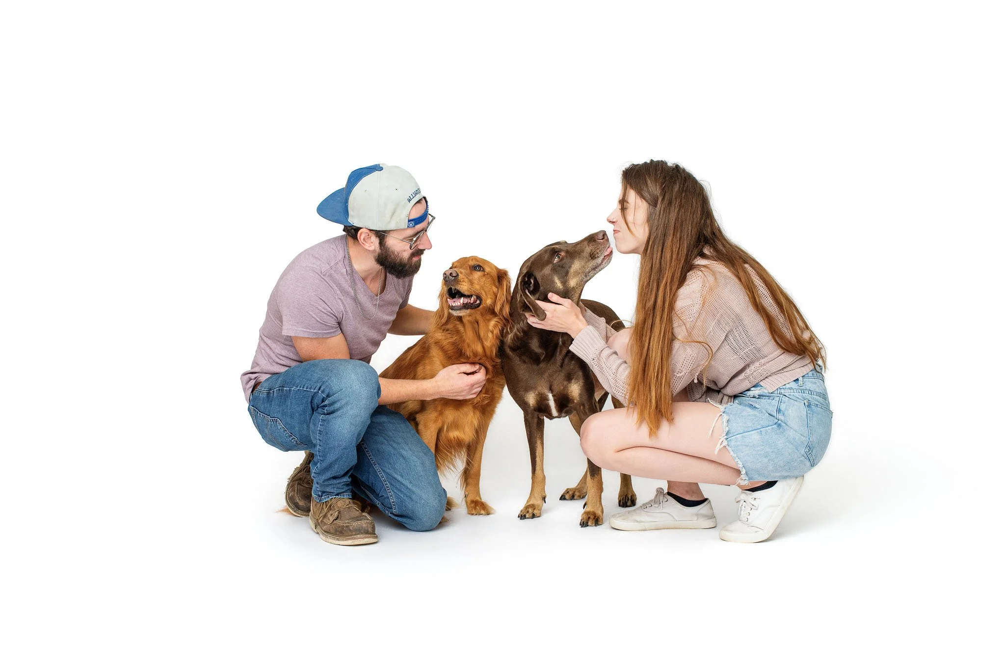 : A couple crouches on a white studio floor with their two dogs, Lilah leaning into the man while Hank stretches up to lick the woman's face.