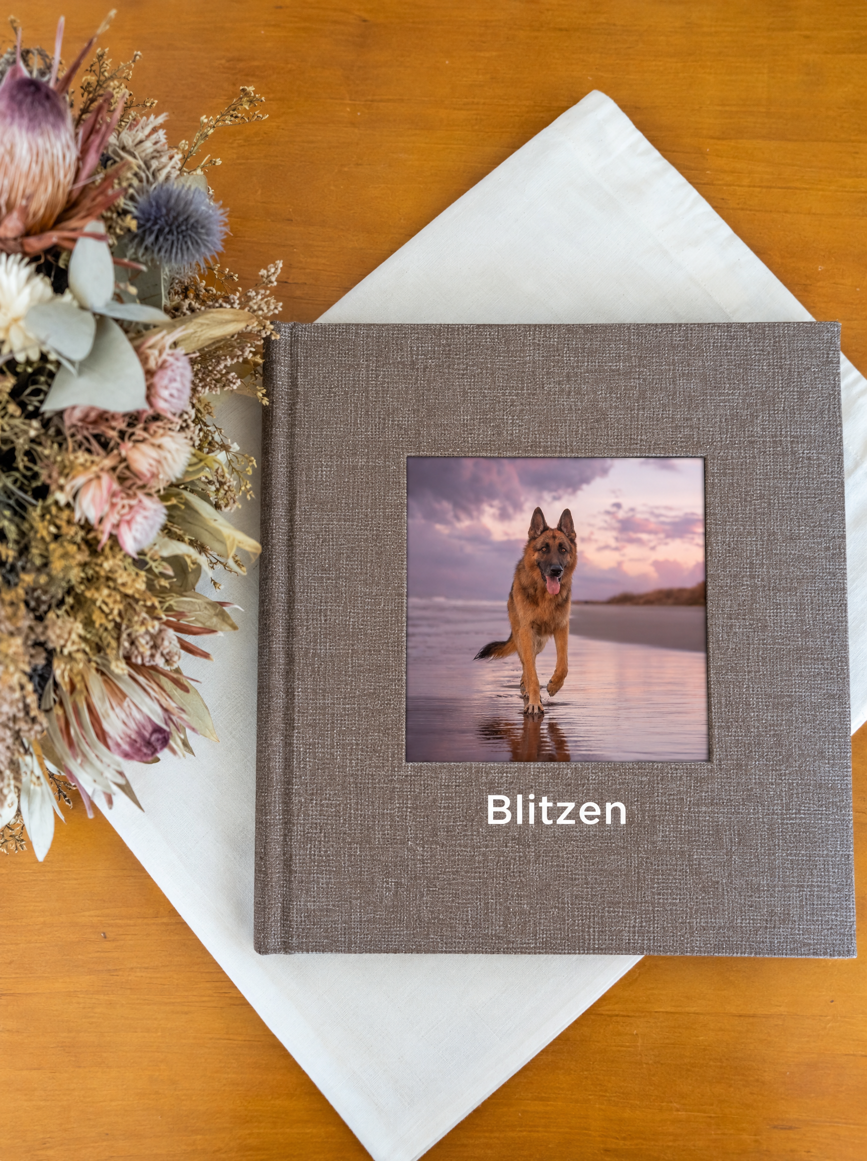 Photo album titled “Blitzen” featuring a German Shepherd running on a beach, surrounded by dried flowers and a white cloth on a wooden table.