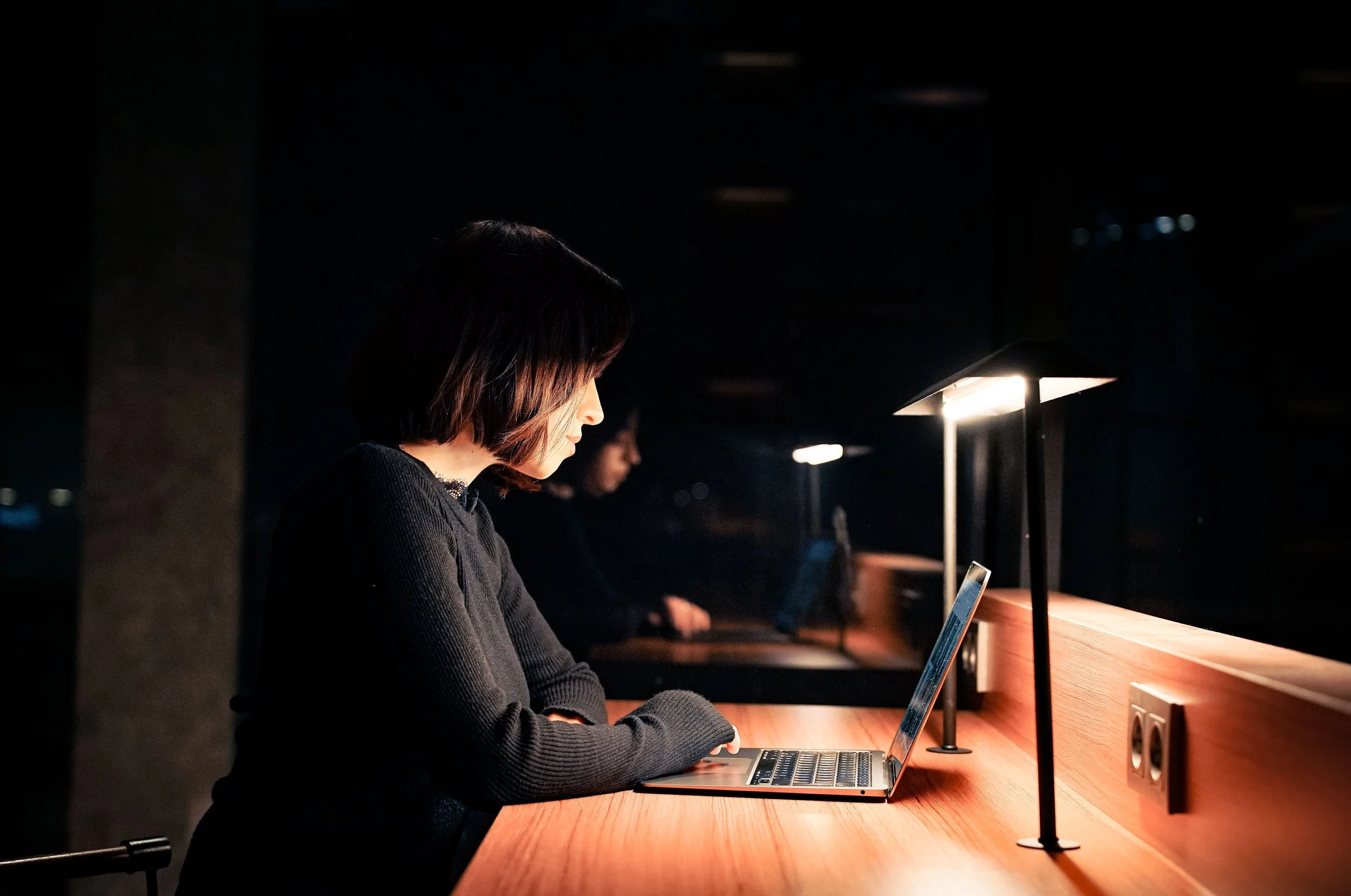 A woman with shoulder-length dark hair sitting at a wooden table in a dimly lit room, looking at her phone while a lamp with a lamp shade provides light.
