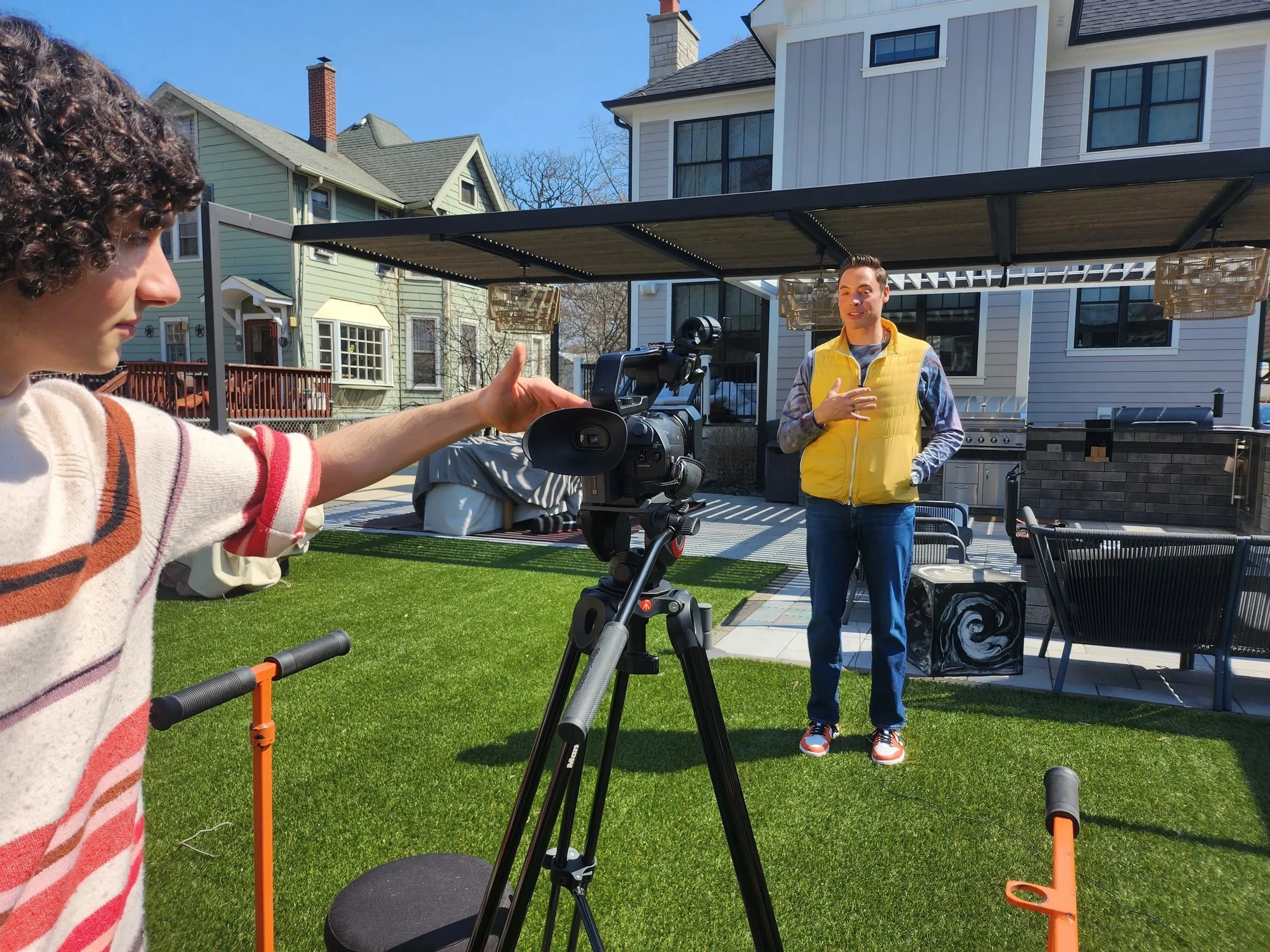 A man in a yellow vest and sneakers is talking during an outdoor interview or recording session. A woman with curly hair and a striped sweater is pointing at a camera, which is set up on a tripod, on a green lawn in front of a modern house. The scene is sunny with a clear blue sky.