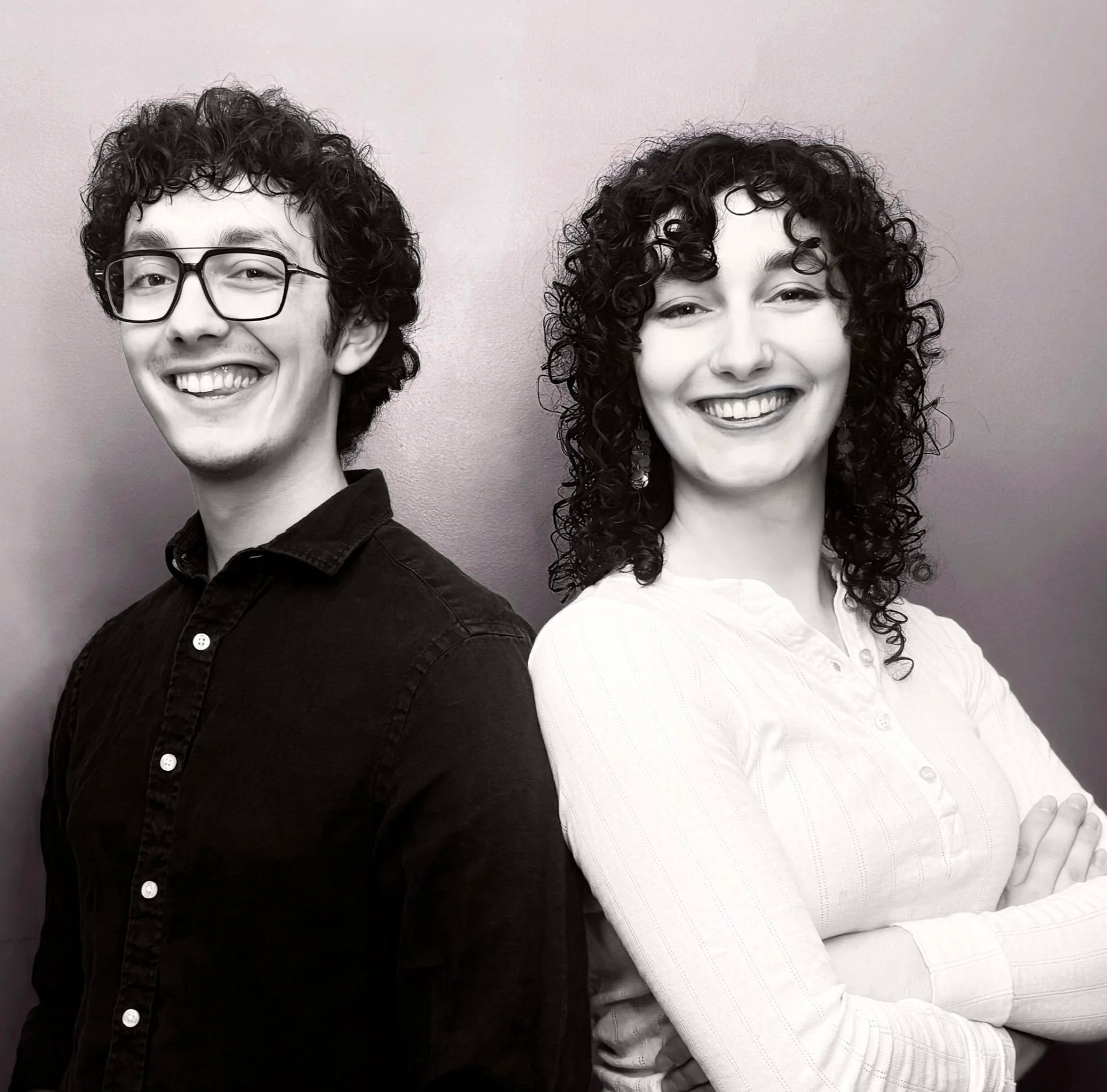 A black and white photo of a young man with glasses and curly hair smiling, standing next to a young woman with curly hair and a smile, both with their arms crossed, against a plain wall.