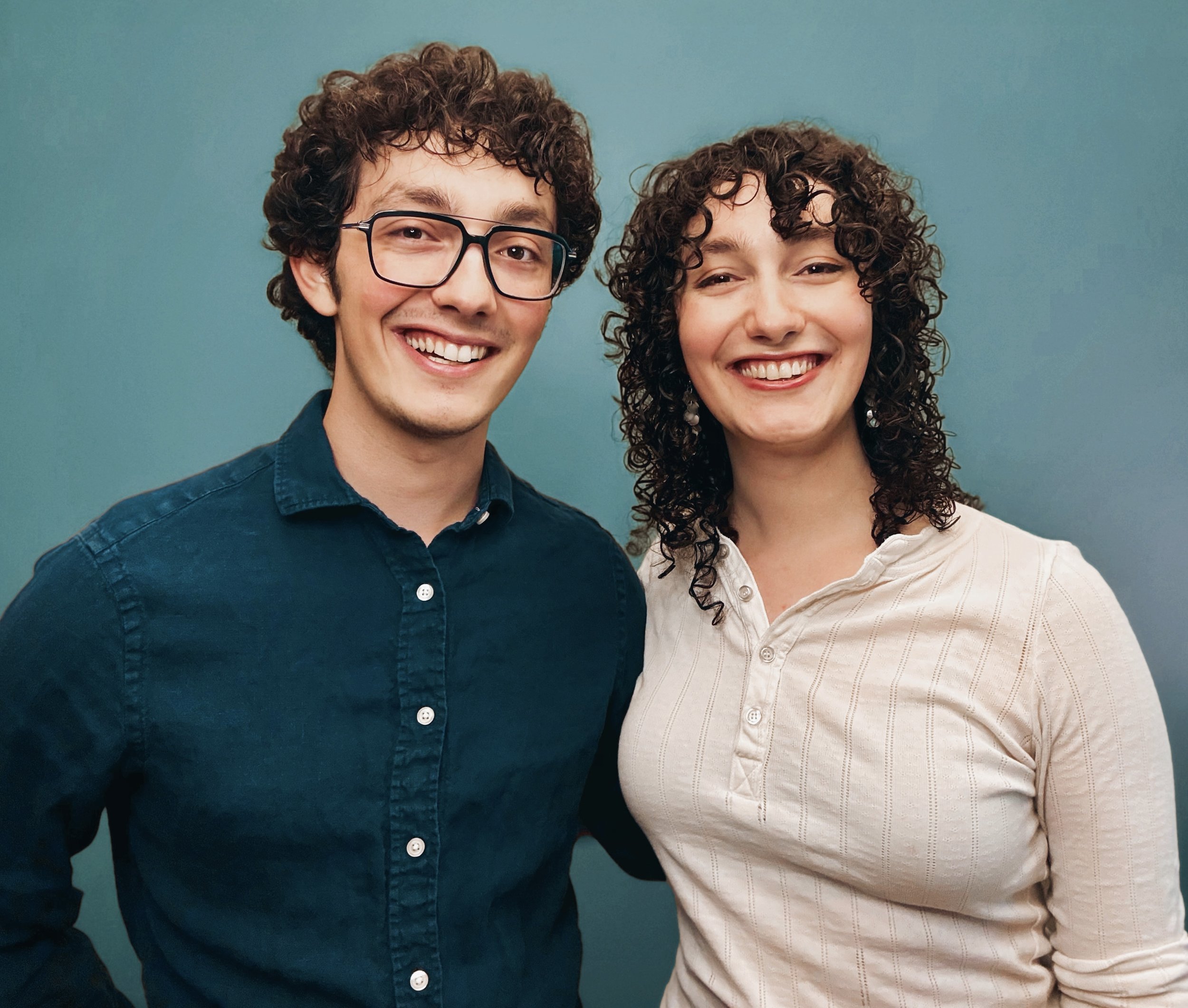 Close-up of smiling man with curly hair, glasses, wearing a dark blue shirt and woman with curly hair and white blouse, both smiling against a teal background.
