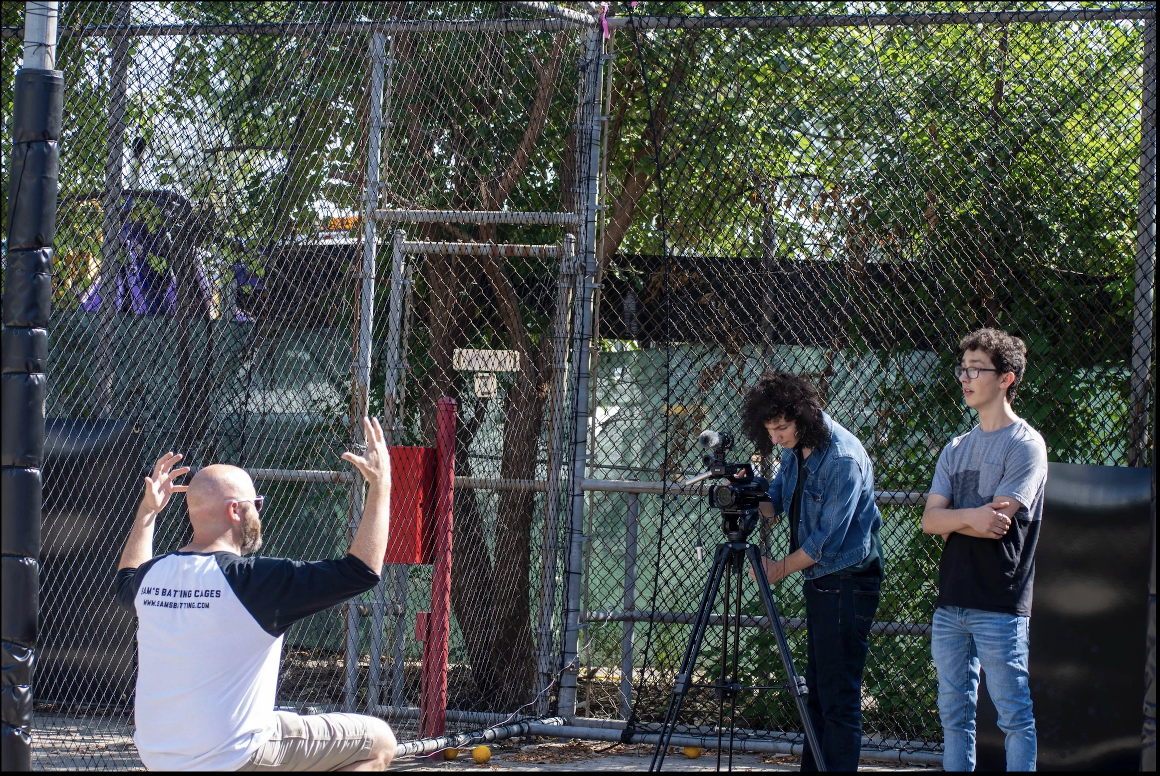 A group of people at an outdoor batting cage facility, with a chain-link fence in the foreground and trees in the background. One person is seated, gesturing with hands, while another is operating a camera and a third is standing nearby with arms crossed.