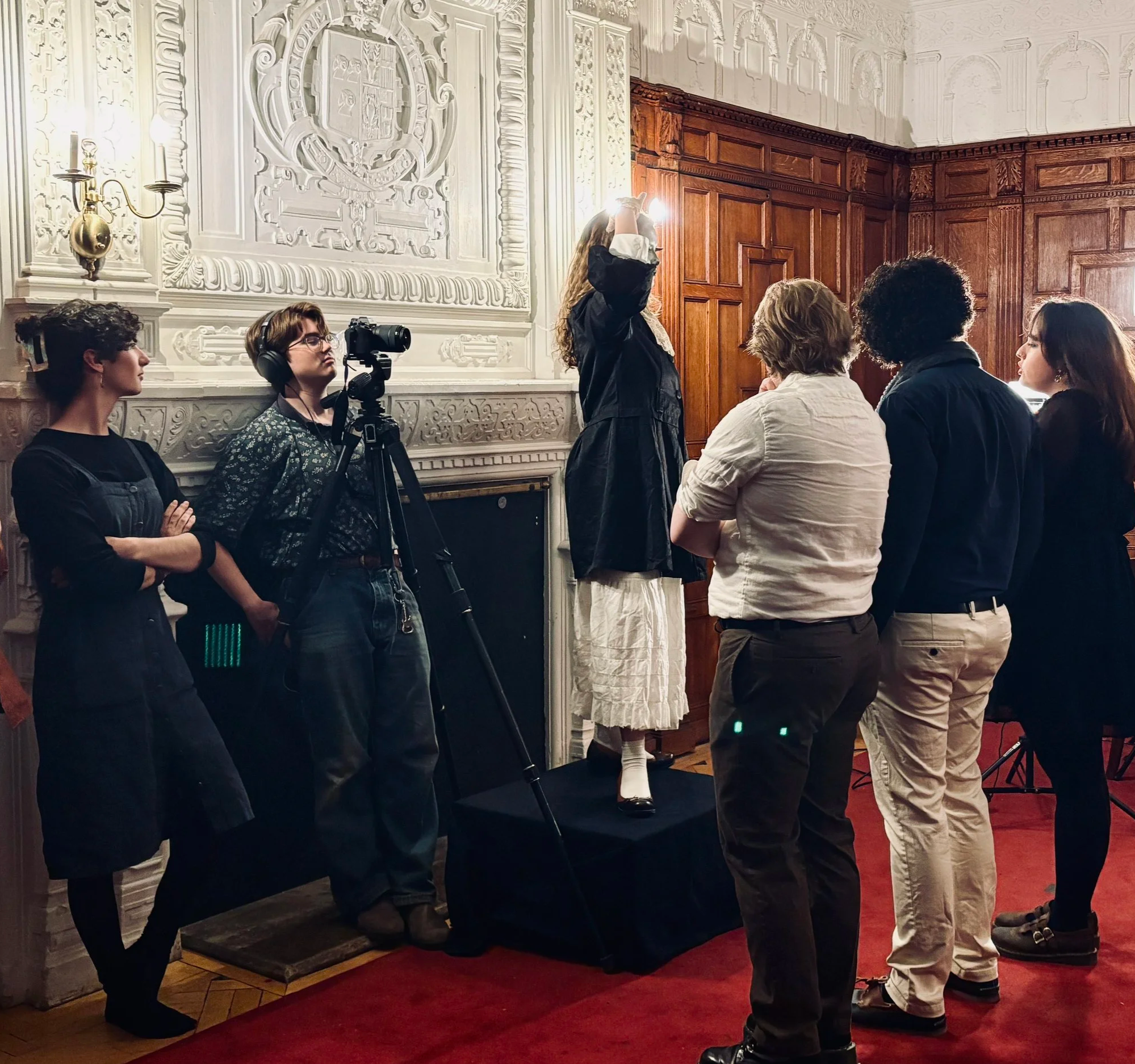 A group of people, including a woman with long hair taking a photo or video, standing in a room with ornate white walls and wooden paneling, and red carpeting.