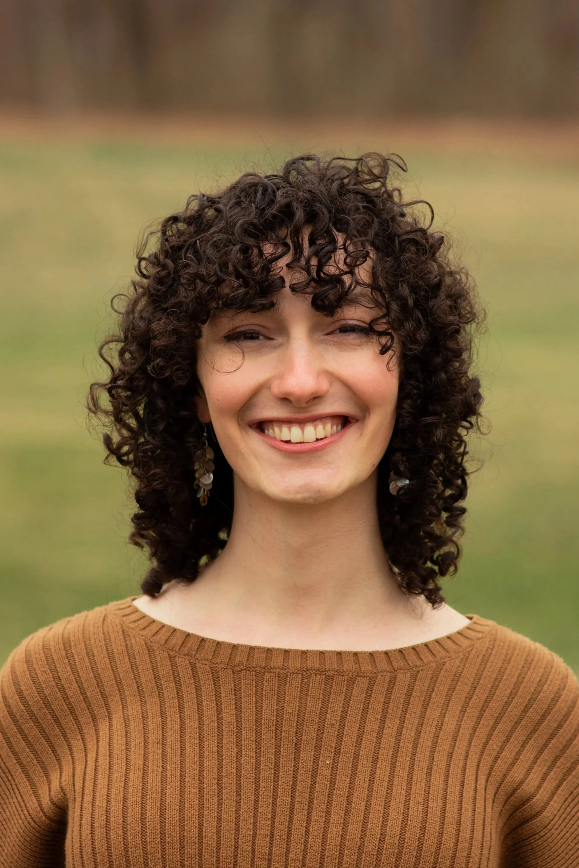 A woman with curly brown hair smiling outdoors, wearing a brown sweater.