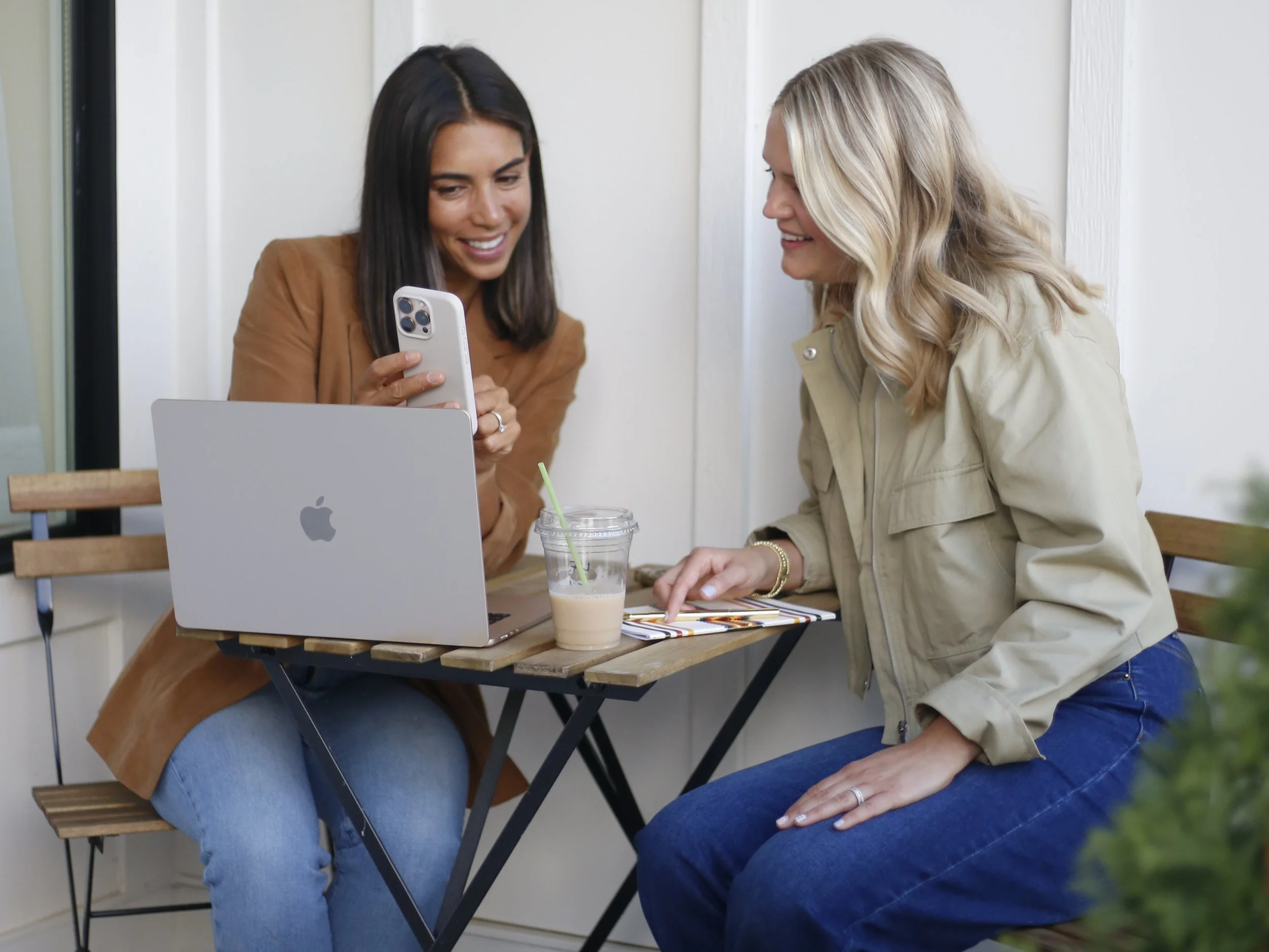Two women sit at a small wooden table outdoors, smiling and looking at a smartphone together. One woman is holding a clear plastic cup with a green straw, likely containing a beverage. A silver laptop with an Apple logo is open on the table.