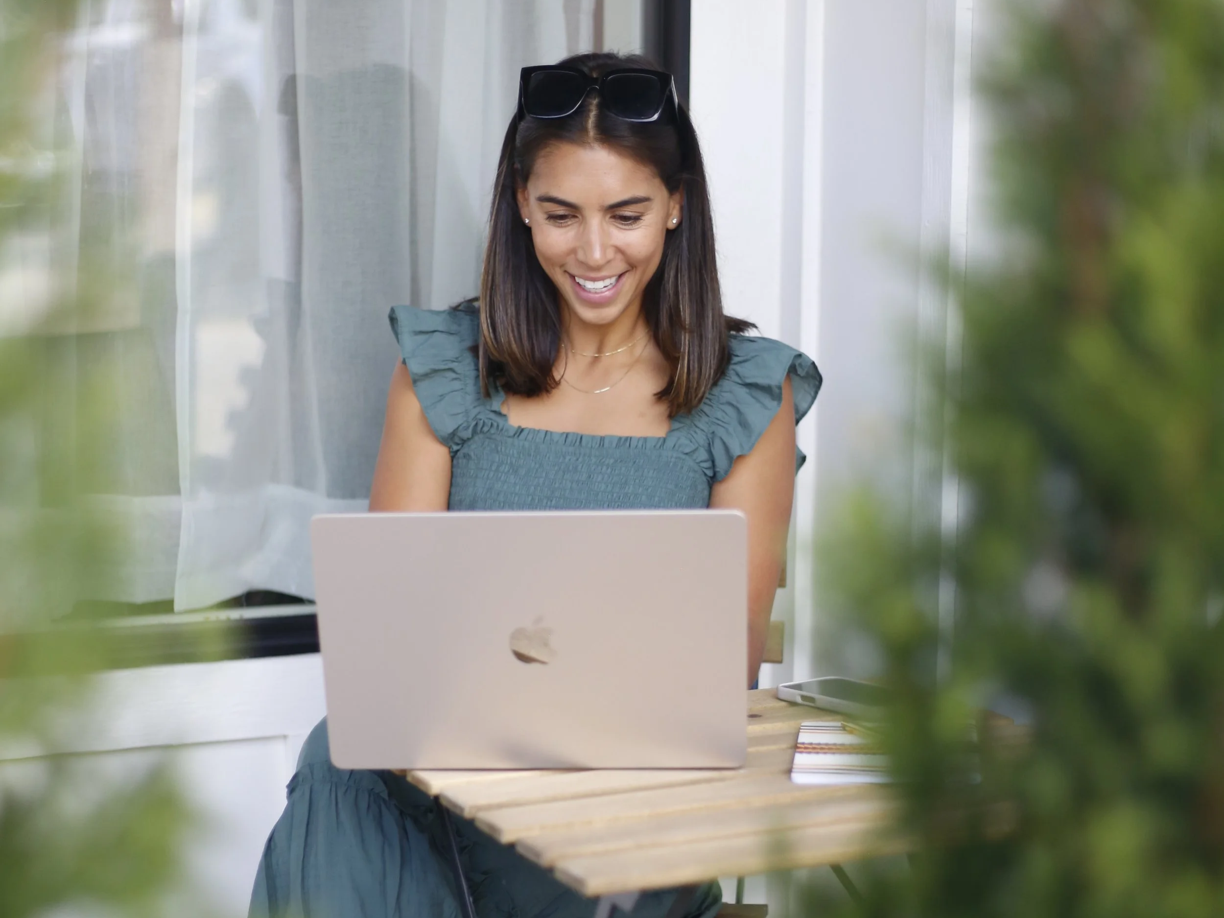 A woman with dark hair, wearing sunglasses on her head and earrings, smiling and looking at her laptop sitting outdoors with green plants surrounding.