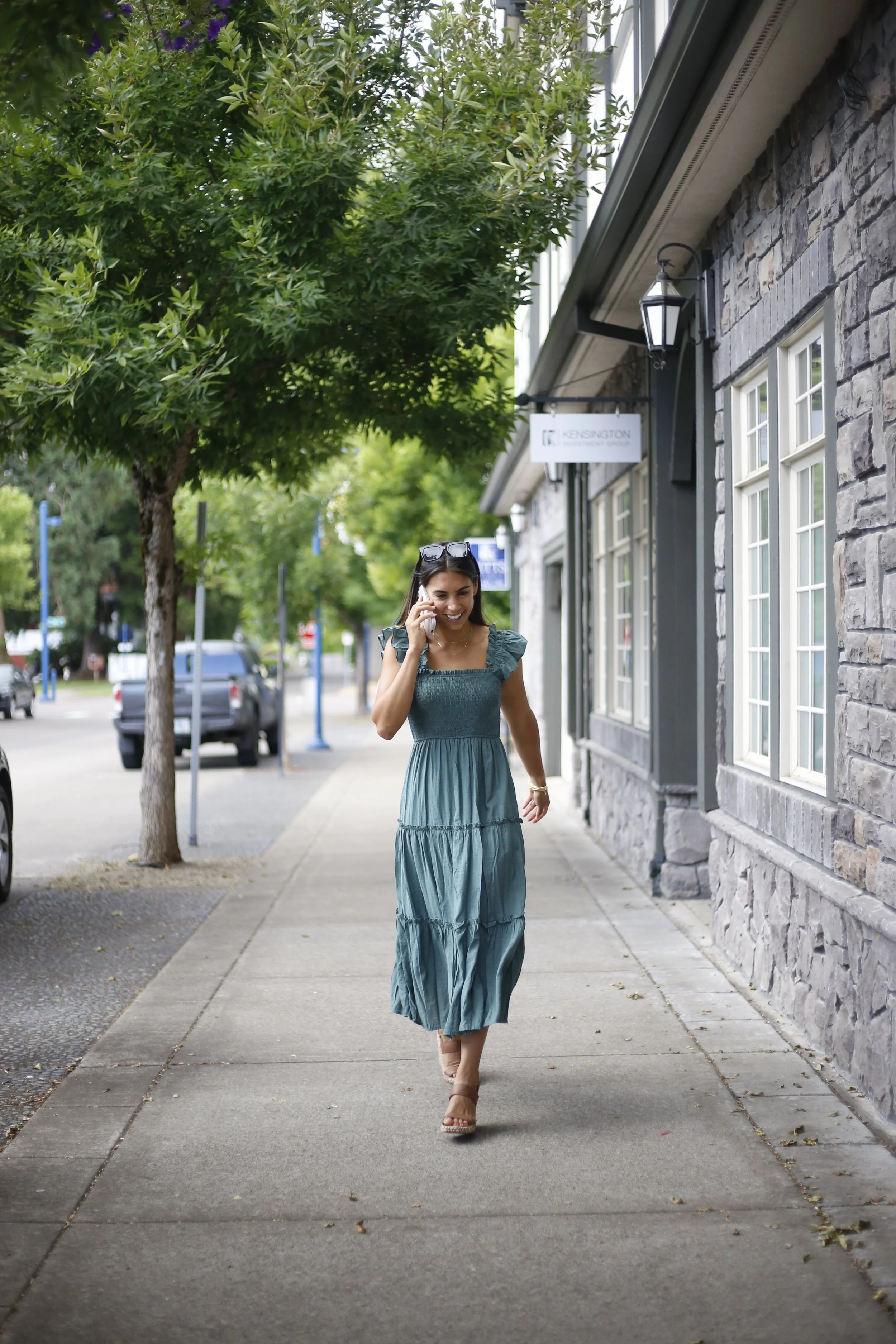 A woman in a teal, layered maxi dress walking down a sidewalk while talking on her cell phone, with sunglasses on her head, next to a stone building and green trees.