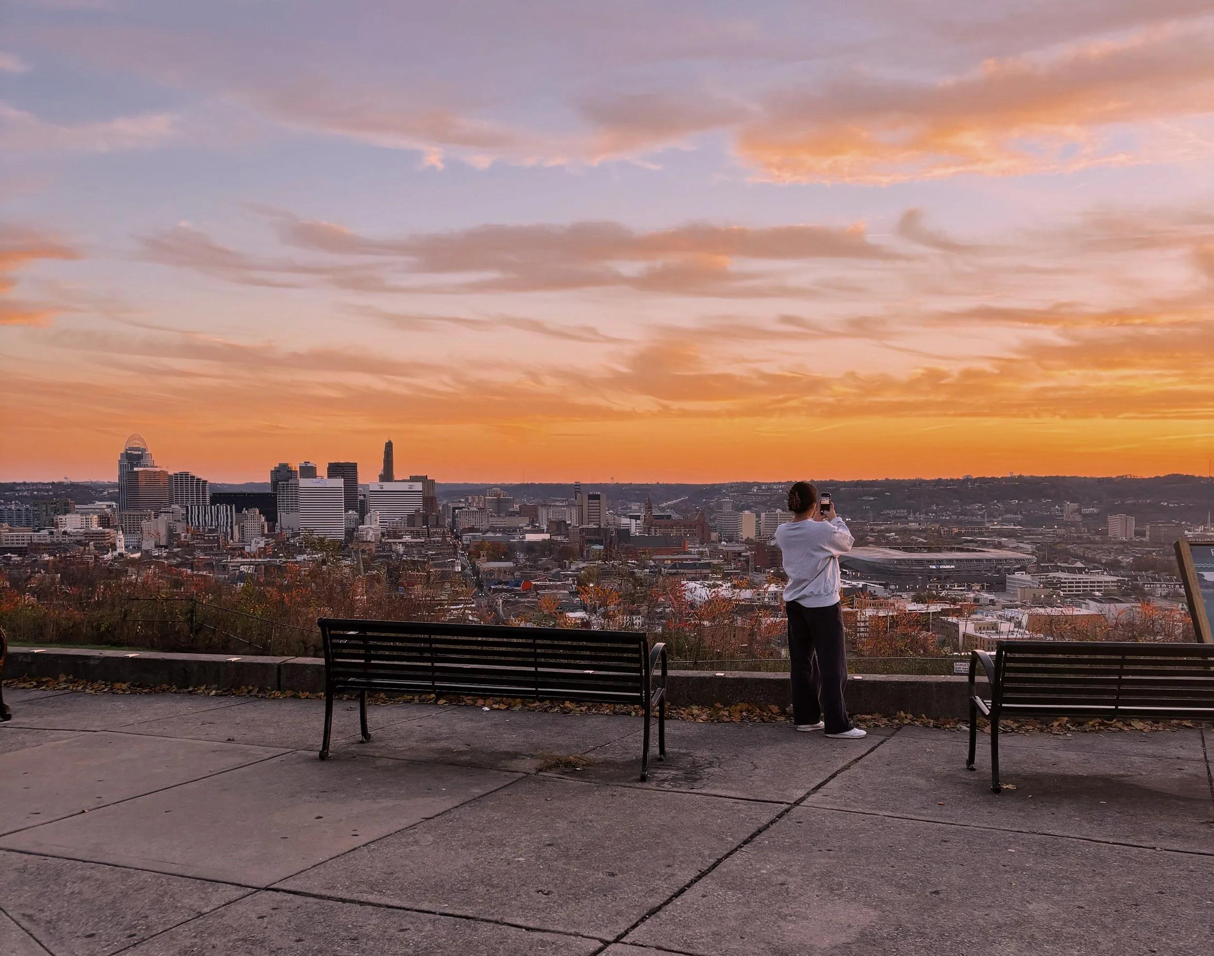Person standing on a park ledge taking a photo of a city skyline at sunset with colorful clouds in the sky.