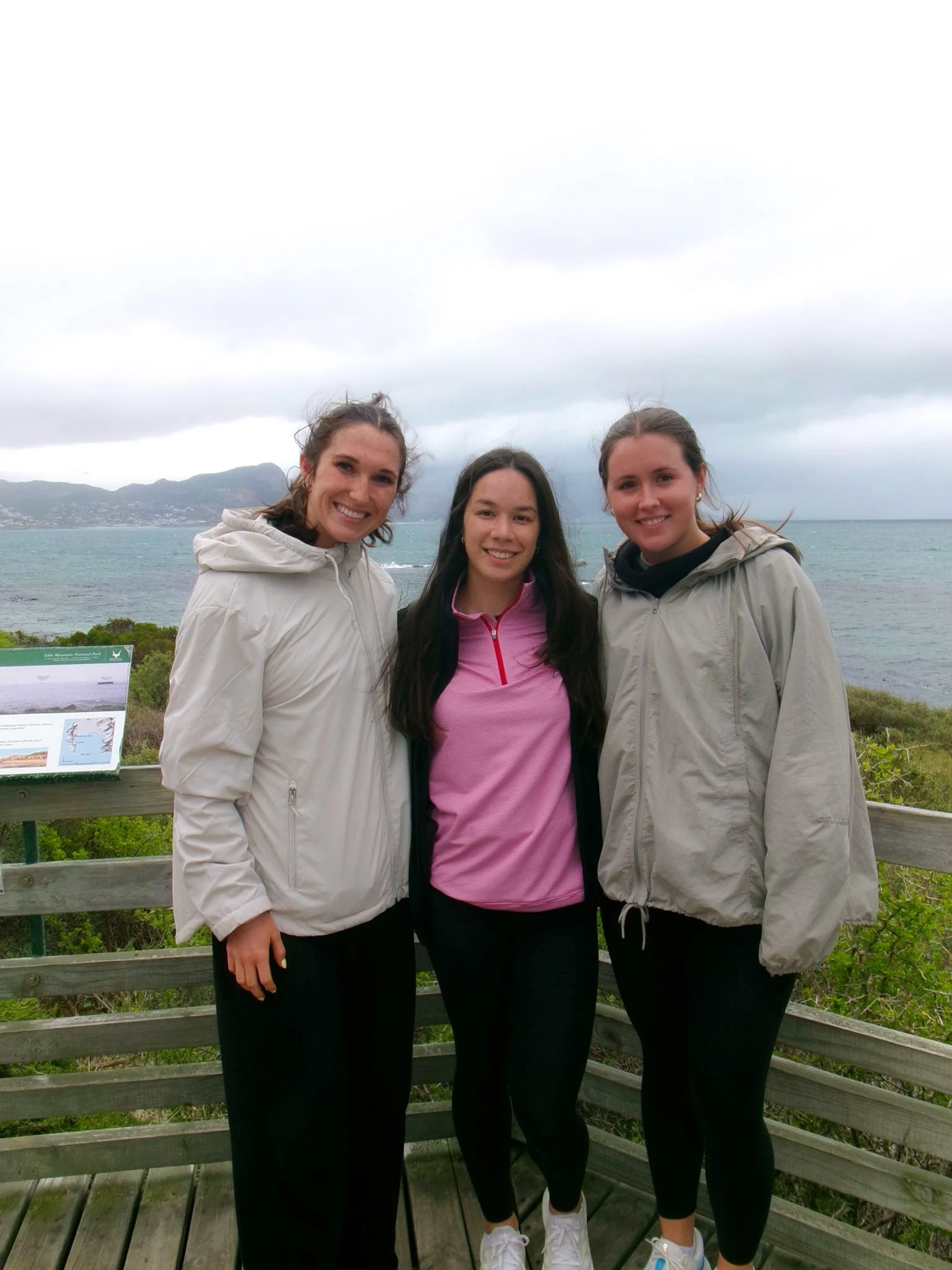Three young women standing close together outdoors near the ocean with mountains in the background, smiling for a photo on a cloudy day.