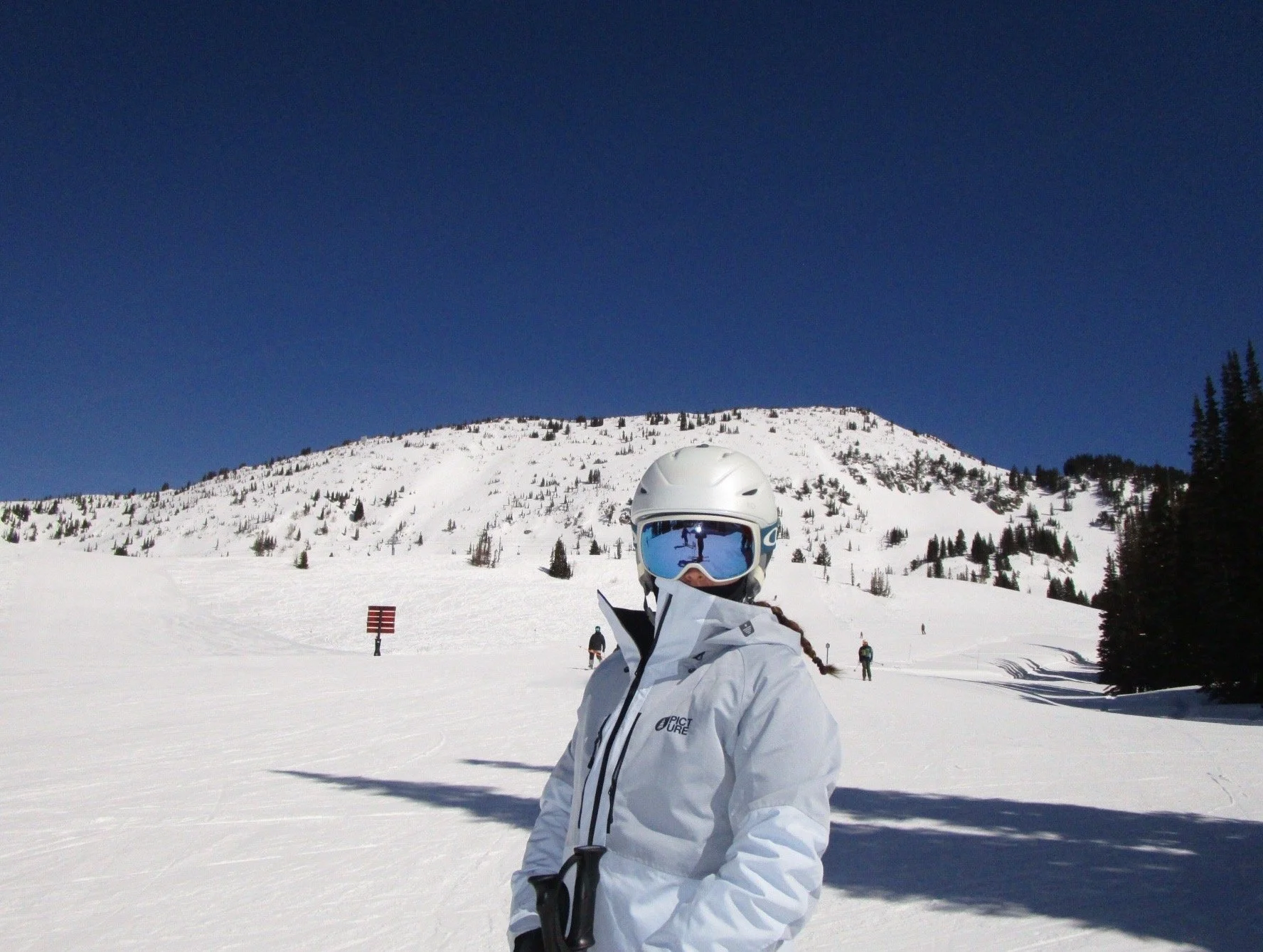 Person dressed in white winter gear and a helmet with ski goggles, standing on snowy mountain slope during a sunny day with a clear blue sky, other skiers in the background, and a snow-covered mountain with trees.