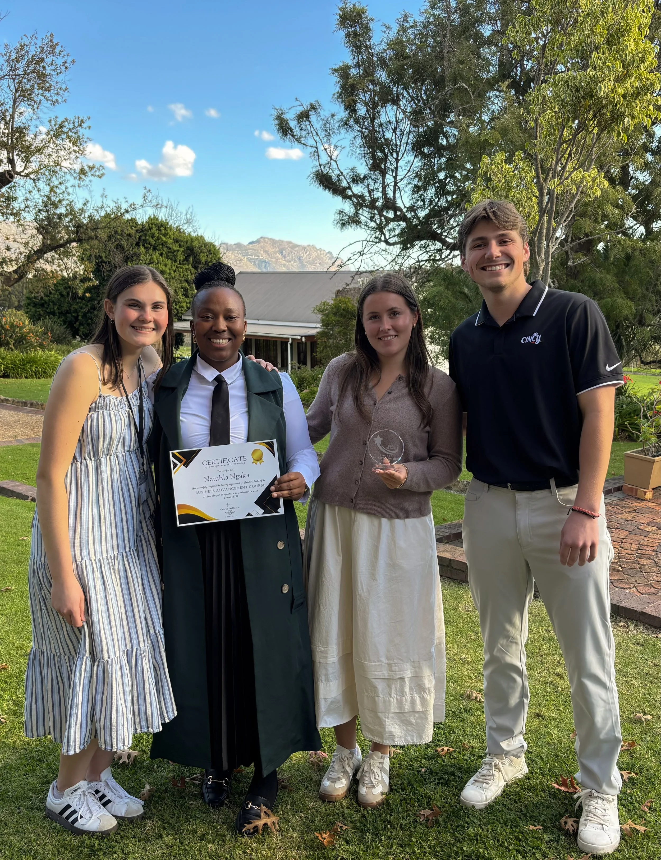 Four young adults smiling and posing outdoors near trees and a house, with one holding a certificate and another holding a glass award, in a setting with mountains in the background.