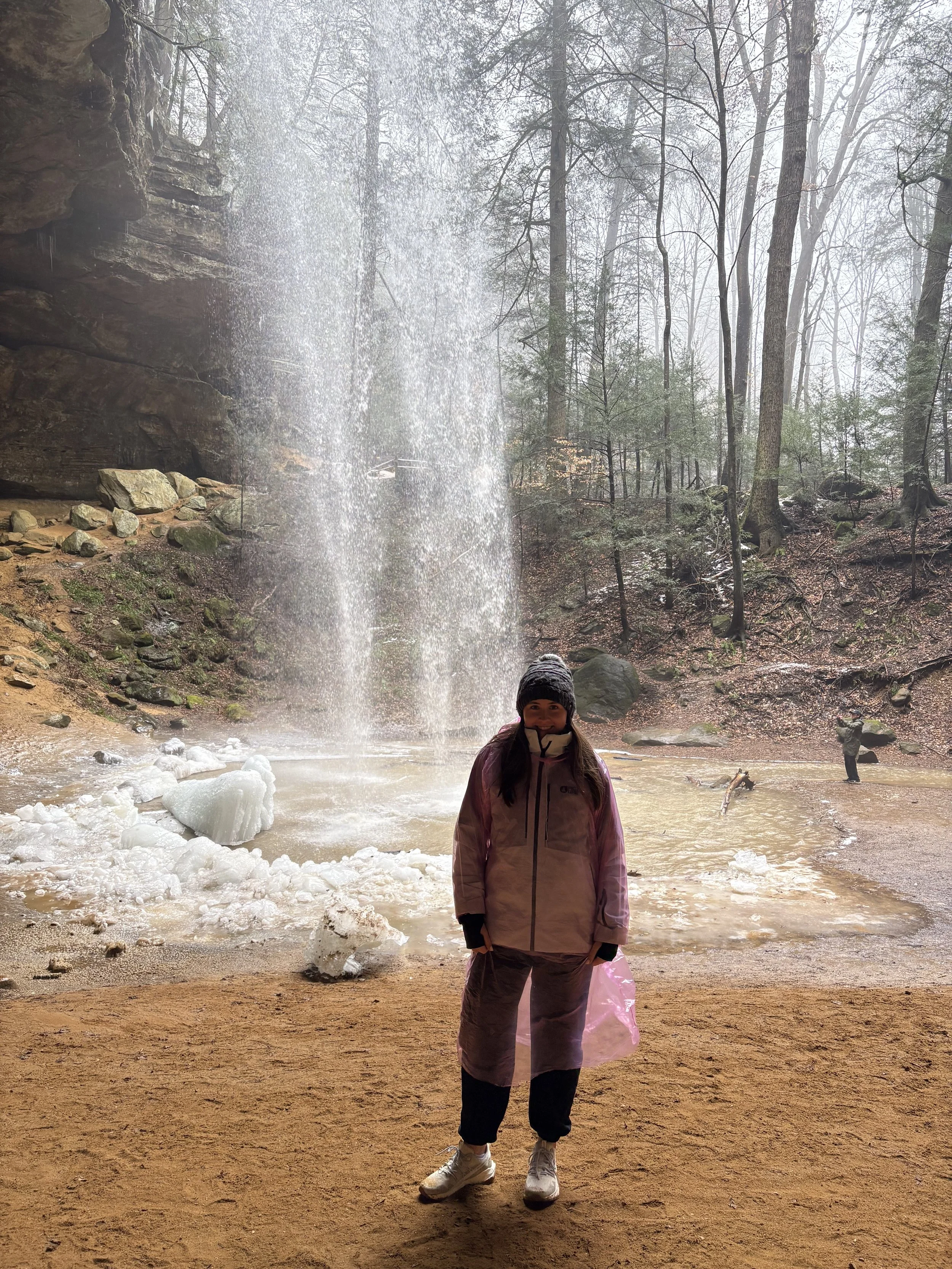 A woman dressed in winter clothing stands on a sandy ground in front of a waterfall in a forest. There are trees and a few people in the background, with snow and ice on the ground.