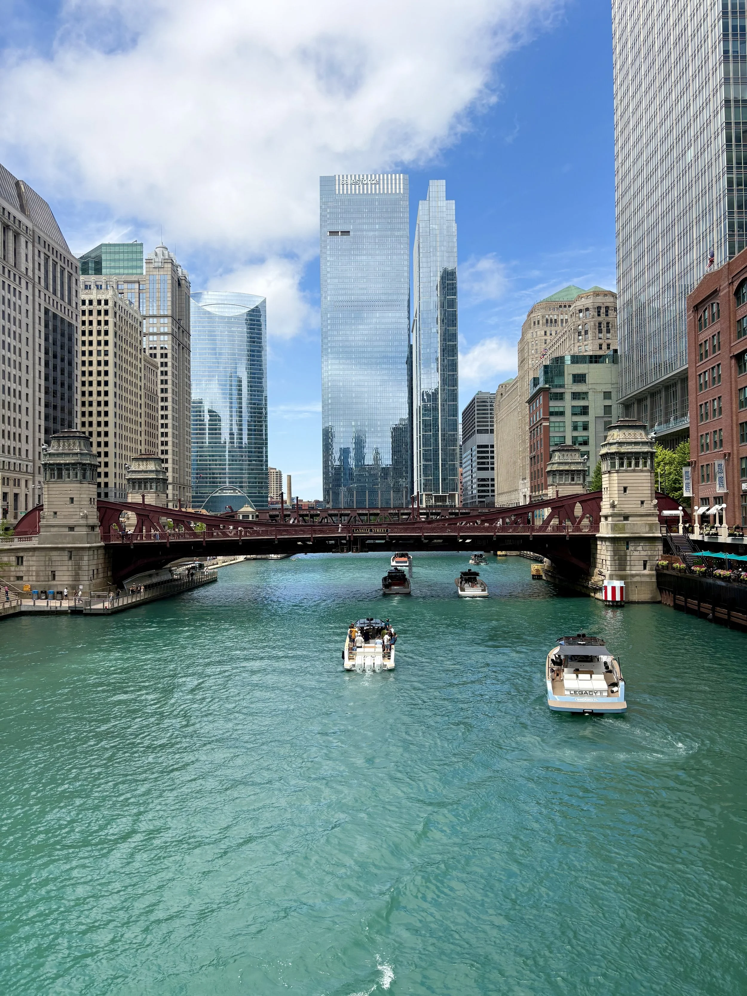 Downtown Chicago skyline with tall glass buildings and boats on the green water of the Chicago River, under a partly cloudy sky.
