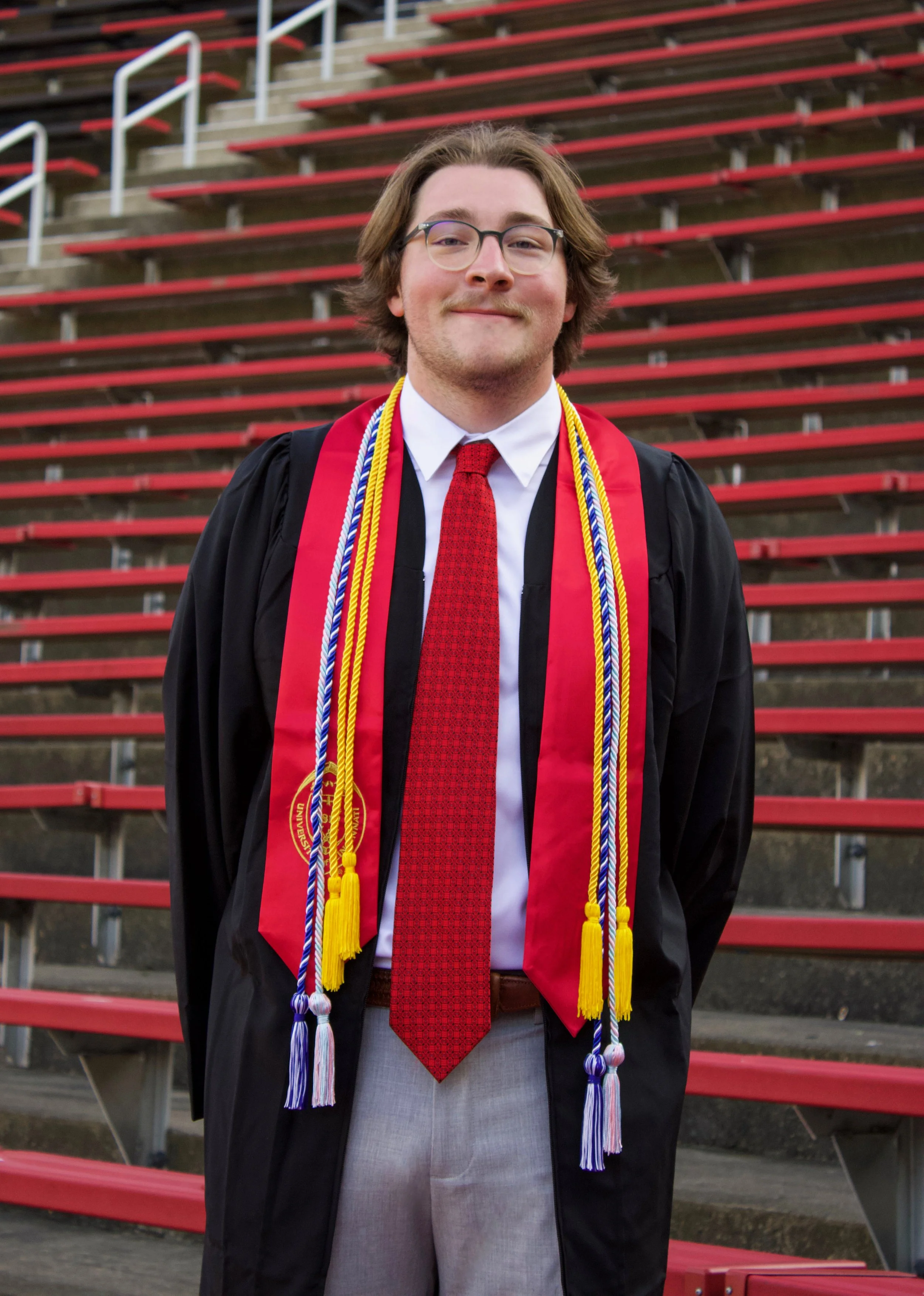 Young man in graduation gown and cap standing on stadium bleachers, smiling, with honor cords and a red stole, wearing glasses and a red tie.