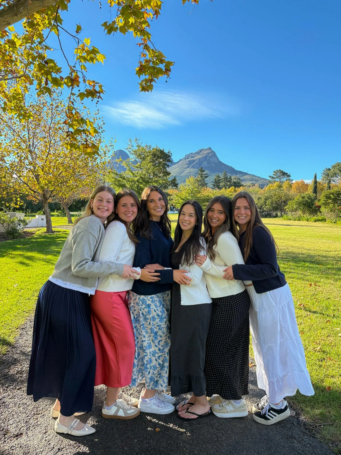Six women smiling and posing outdoors in a park with trees and mountains in the background on a sunny day.