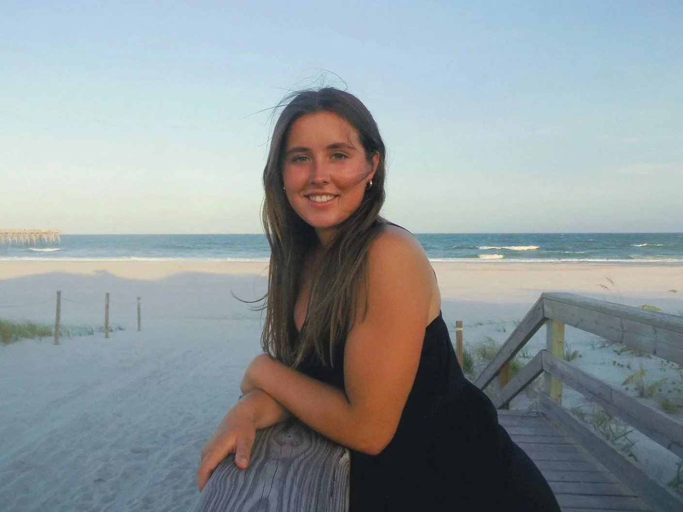 A woman with long brown hair smiling and leaning on a wooden railing at the beach during sunset, with the ocean waves and sky in the background.