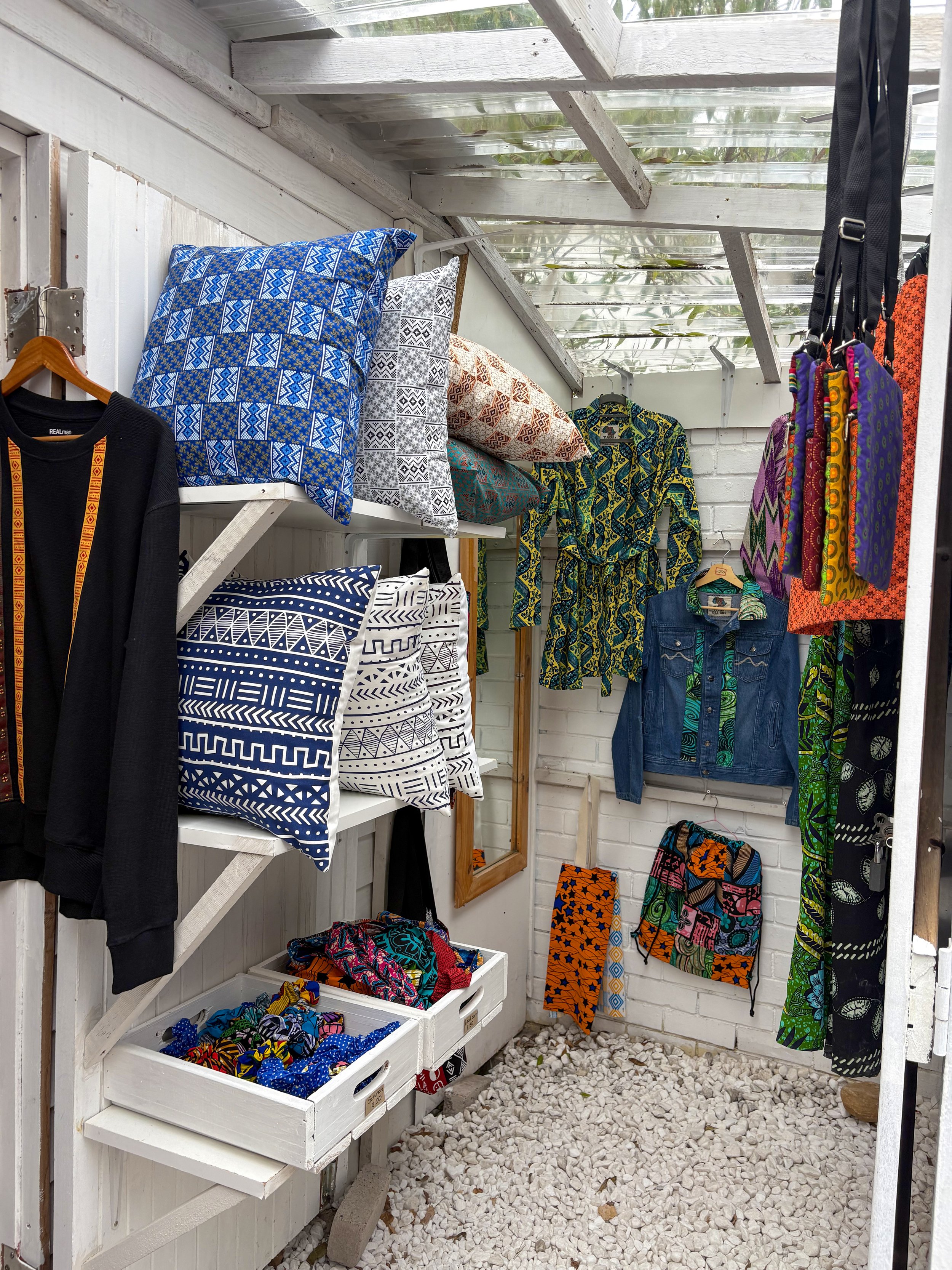 Clothing and accessories display in a small outdoor shop with a white painted wooden wall, shelves, and a stone gravel ground. Items include pillows, shirts, pants, and headbands with vibrant patterns.
