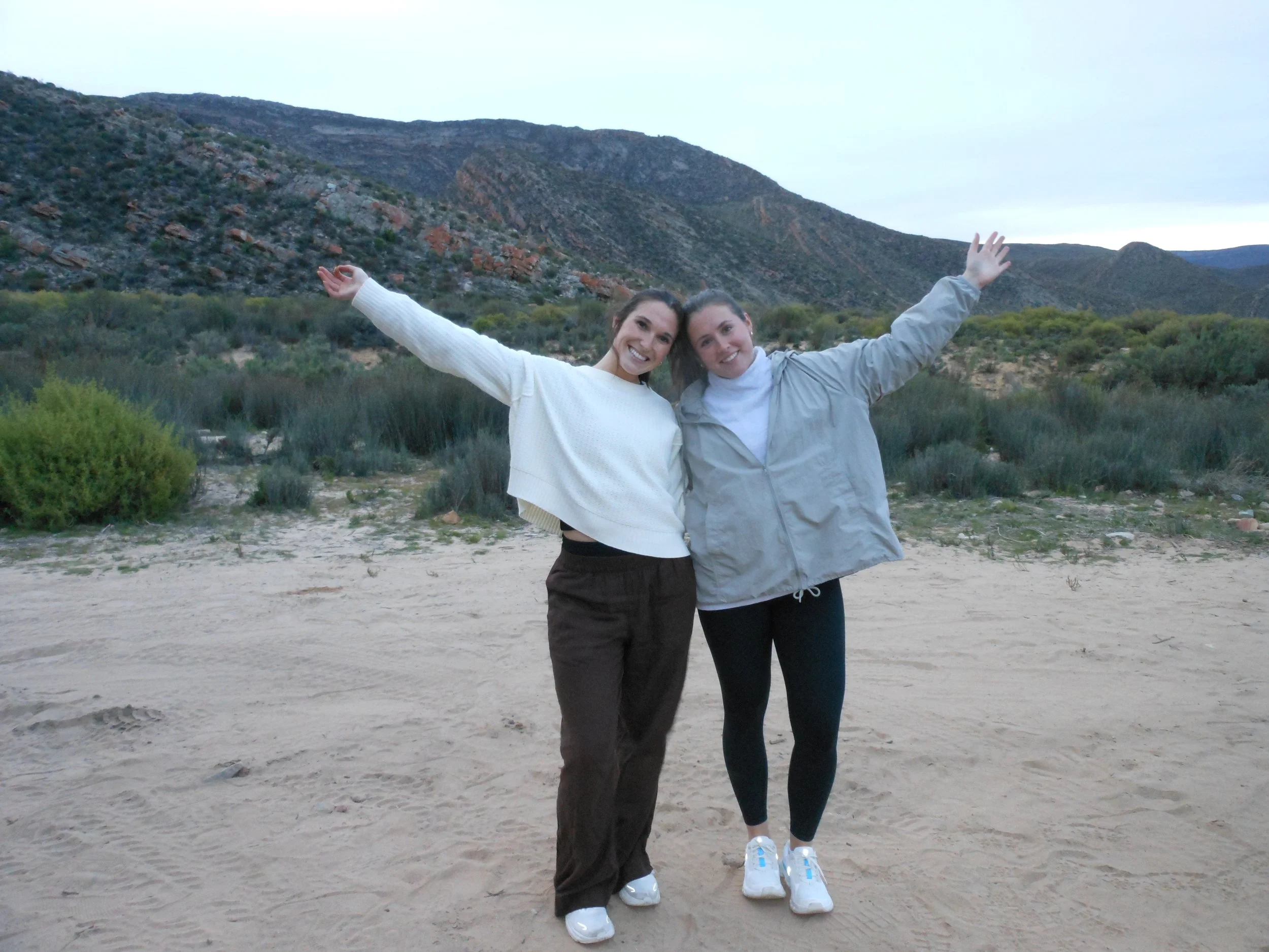 Two women standing on sandy ground with arms outstretched, smiling, with a backdrop of hills and overcast sky.