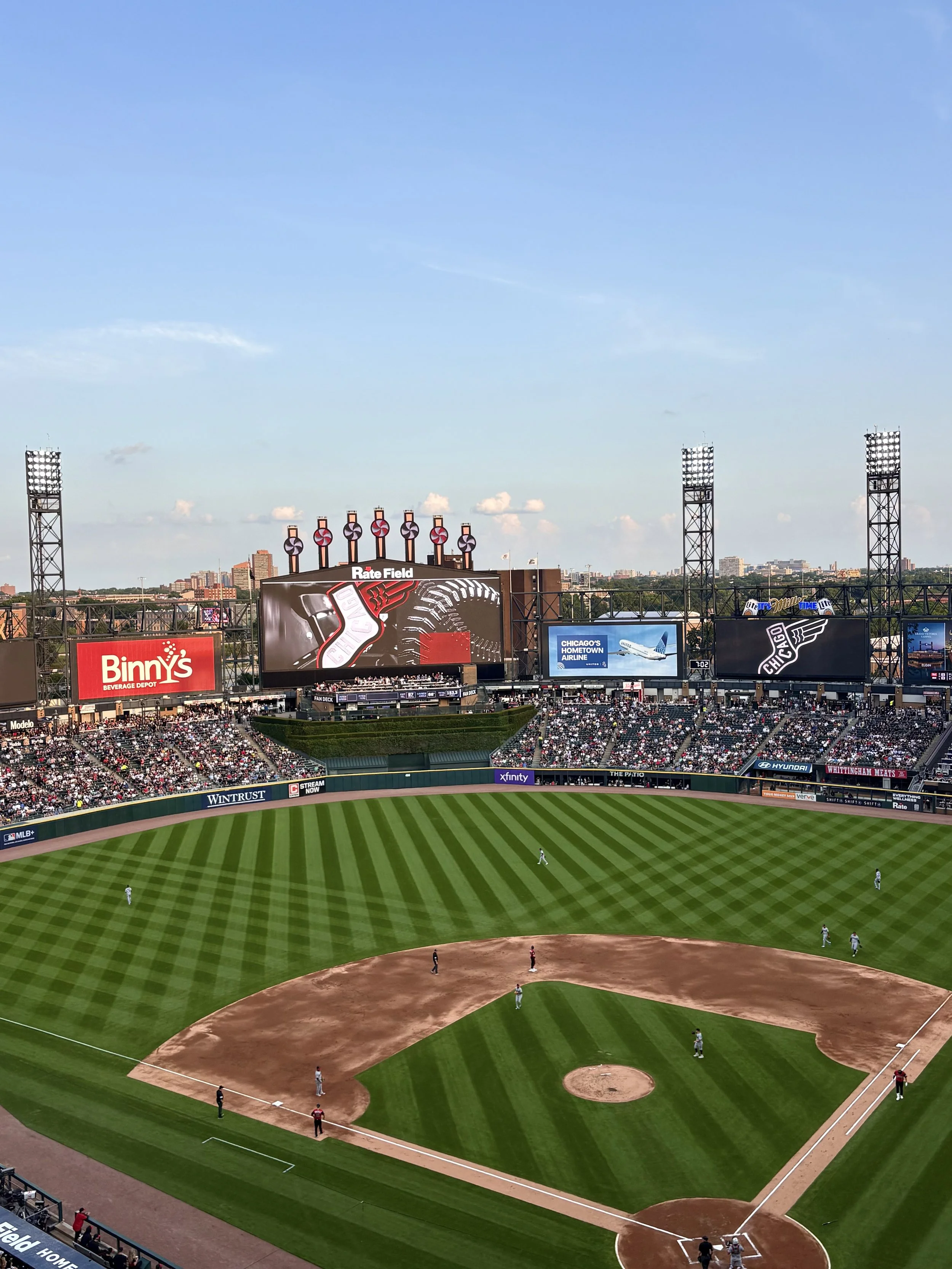 View of a baseball stadium showing the field from the outfield, with players positioned on the field, large electronic scoreboard, advertisements, and spectators in the stands under a clear sky.