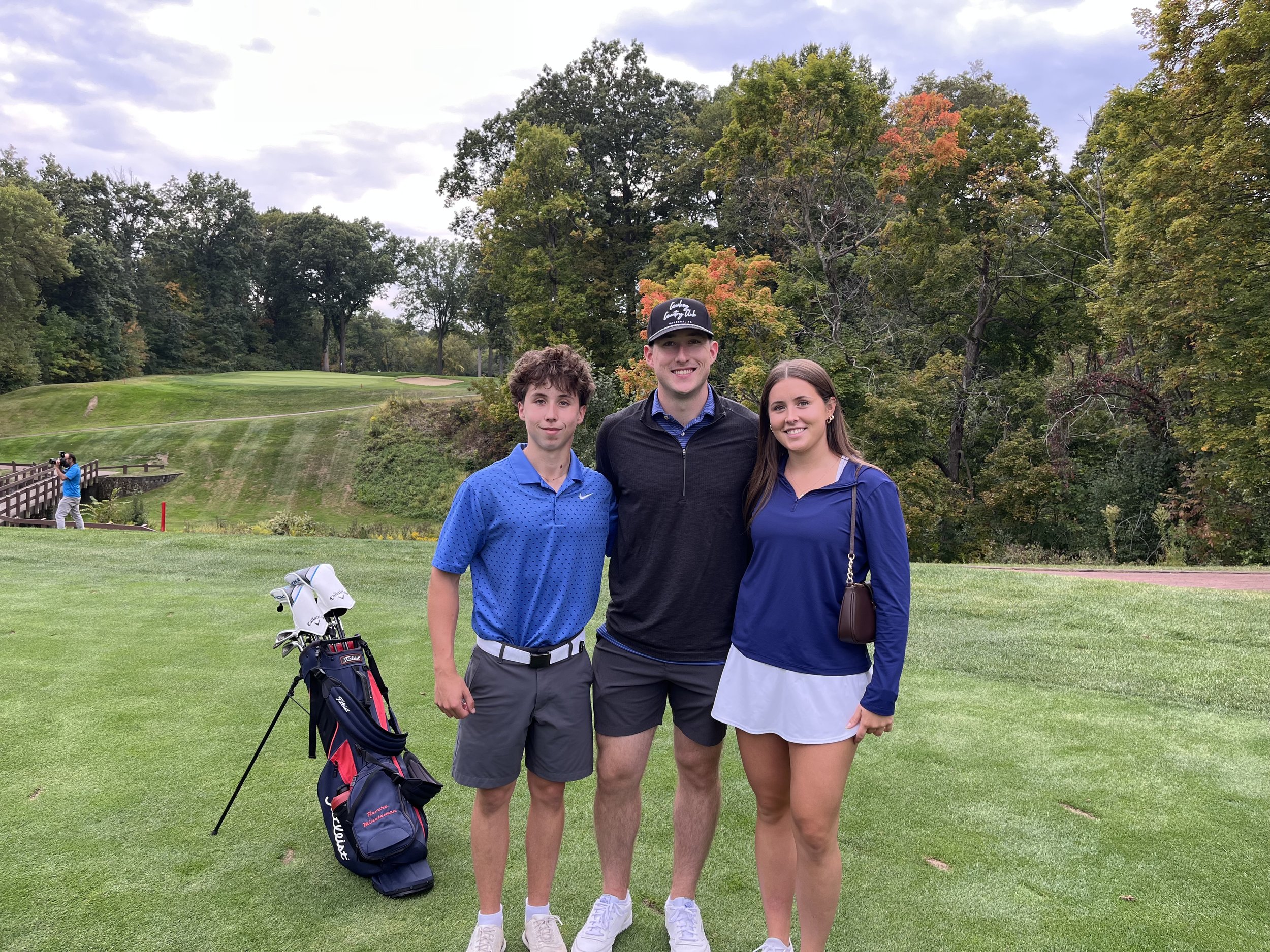 Three young adults standing on a golf course with trees in the background. A person is taking a photo in the distance, and a golf bag with clubs is on the ground in front of the group.