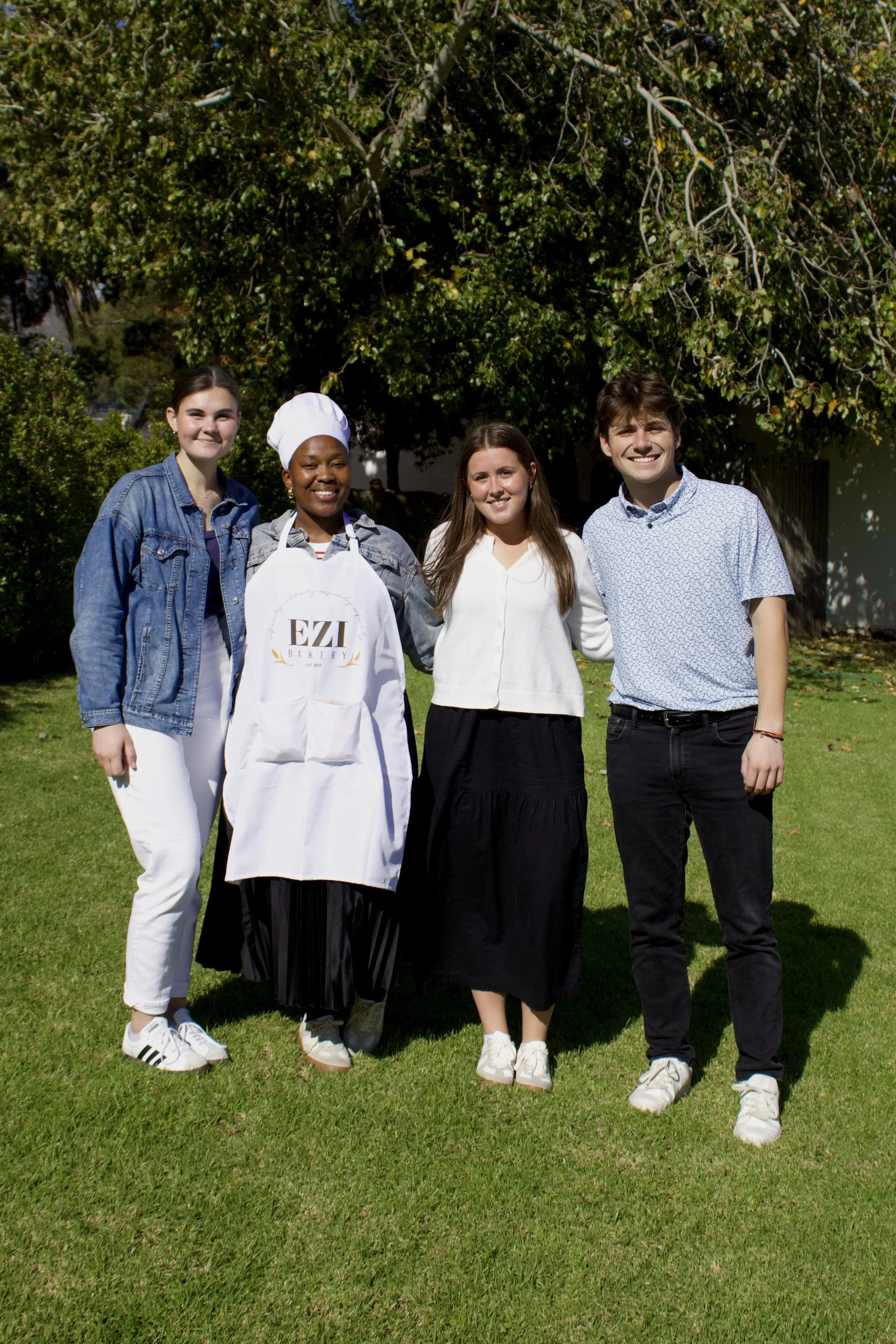 Group of four young people standing on grass in front of a large tree, smiling at the camera. One person is wearing a chef's hat and apron with 'EZI Bakery' logo.