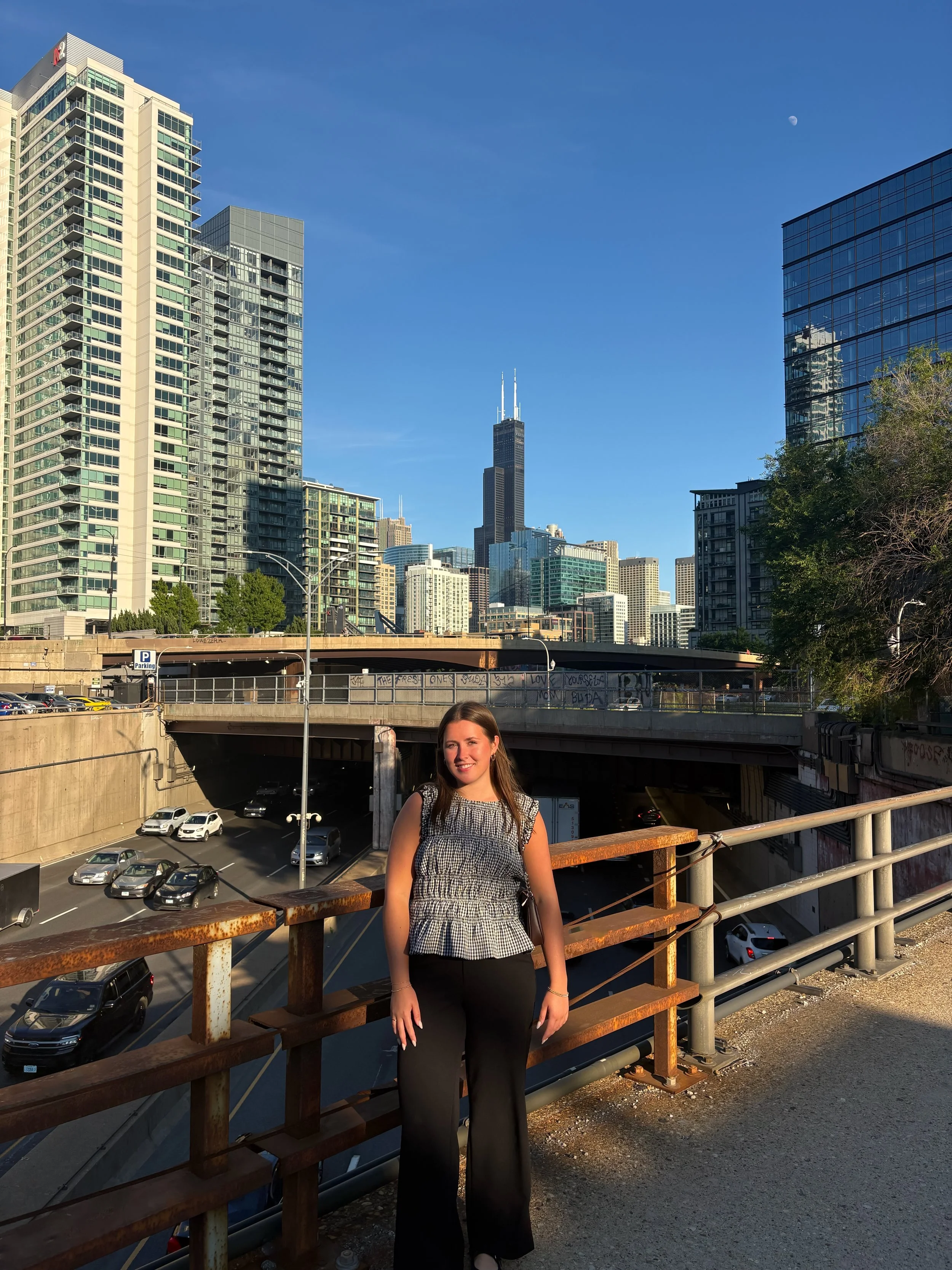 A woman standing on an urban bridge with city skyscrapers in the background, including a tall black building with antennas, a parking lot below, and clear blue sky with the moon visible.