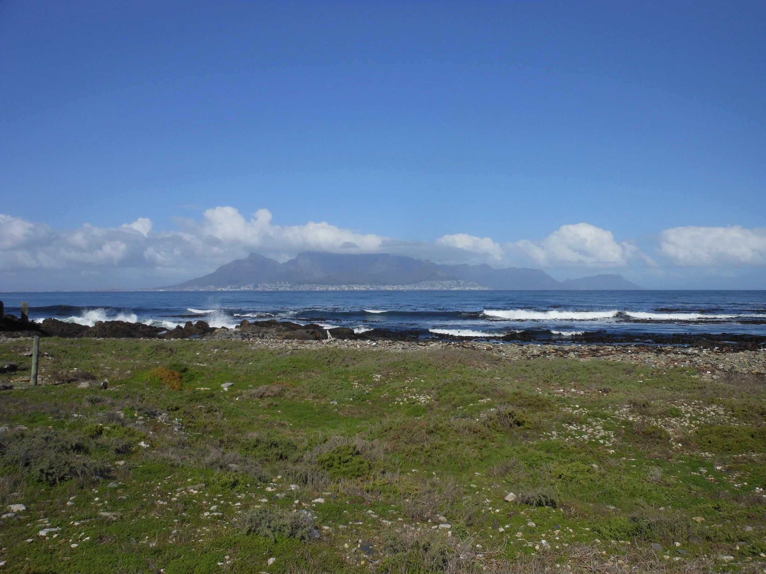 A scenic view of the ocean with waves crashing against the rocky shoreline in the foreground, and a distant mountain range under a blue sky with some white clouds.