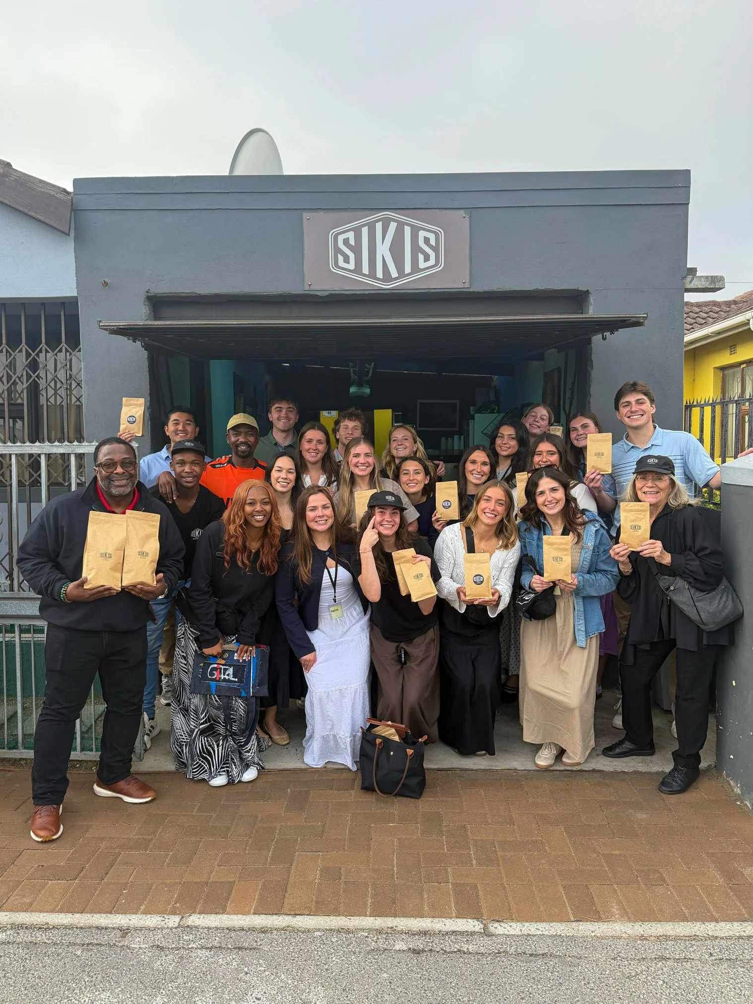 A large group of people, diverse in age and ethnicity, standing in front of a building with a sign labeled 'SIKIS'. Many are smiling and holding brown paper bags, some wearing black hats with the logo. The group appears to be posing for a group photo