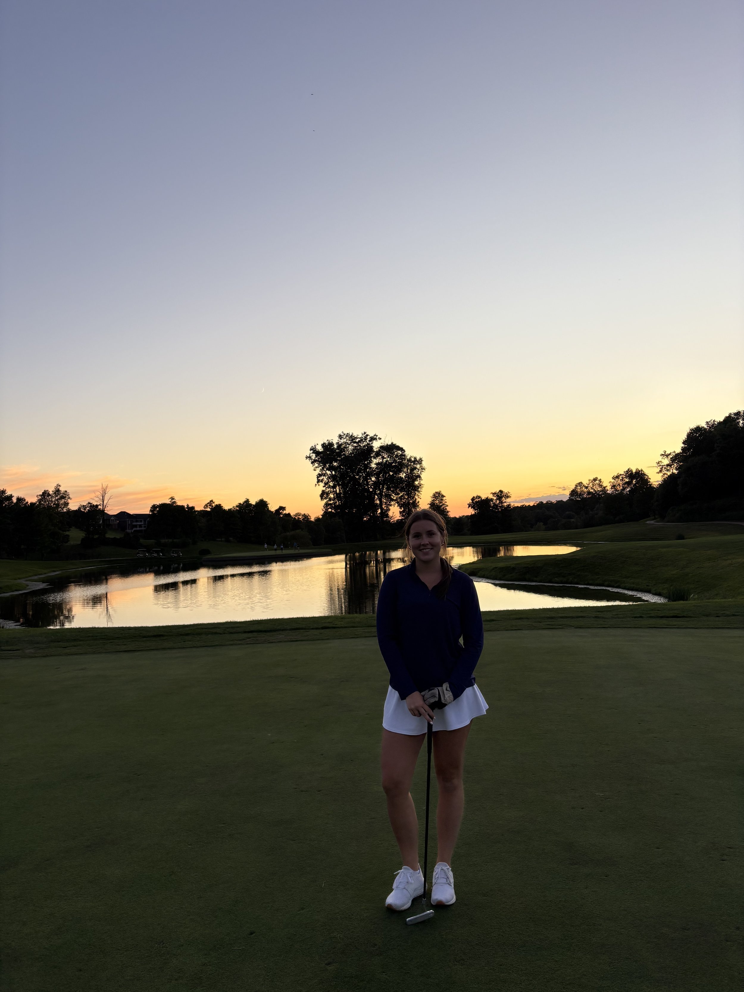 Young woman standing on a golf course holding a golf club, with a pond and trees in the background at sunset.