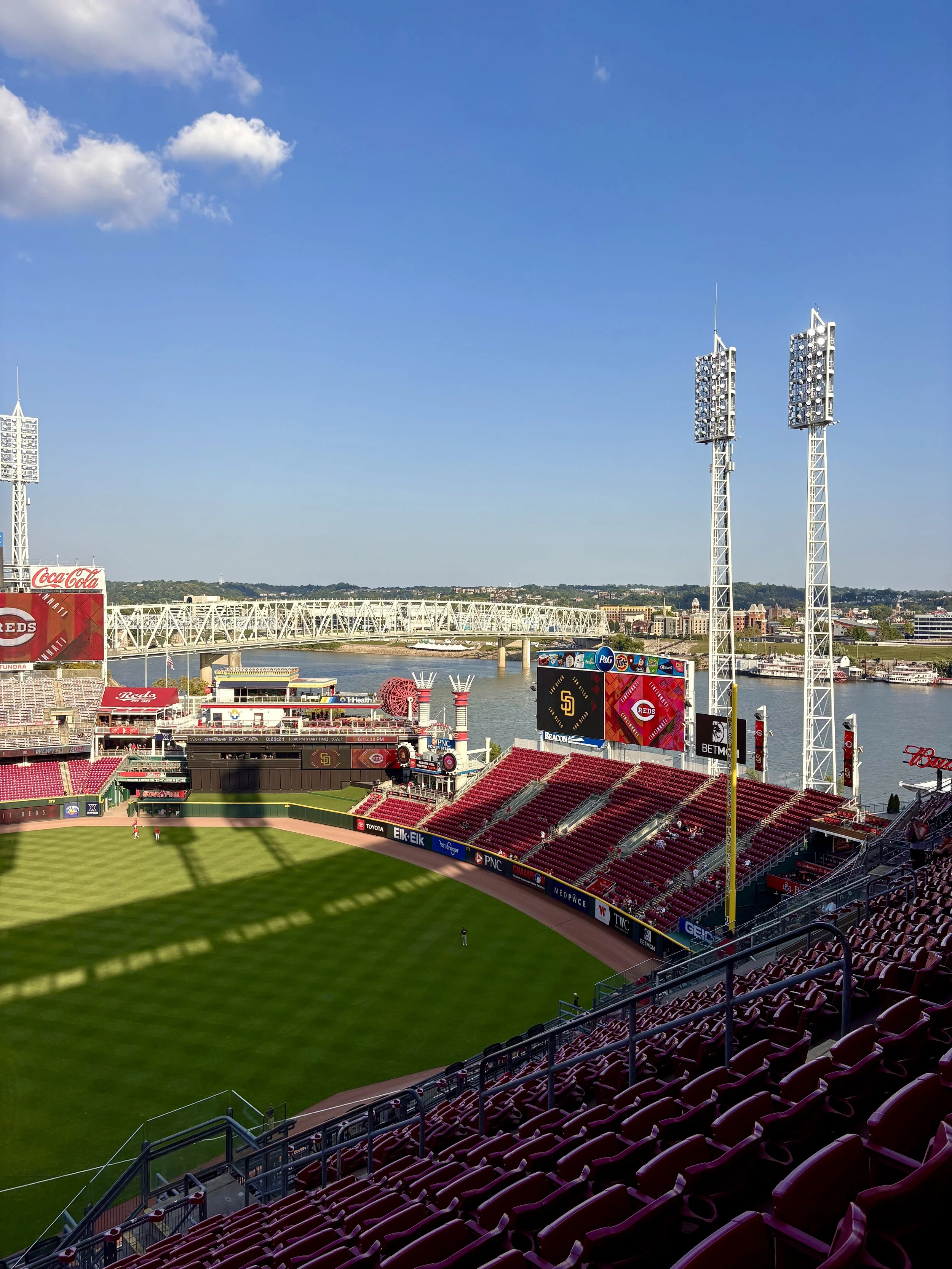 View of a baseball stadium with green field, red seats, and a scoreboard showing team logos, overlooking a river and cityscape under a blue sky with a few clouds.