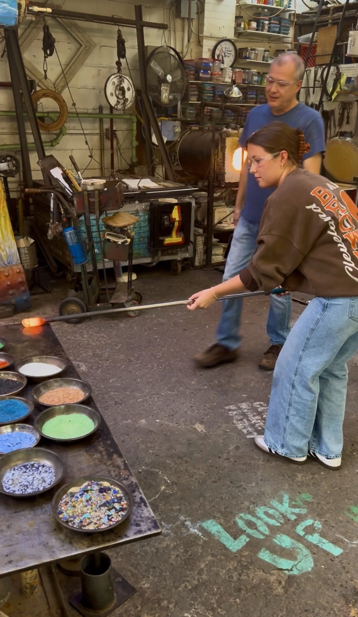 Two women and a man are in a glassblowing workshop. One woman is holding a long rod with hot glass at the end, shaping it. The man is observing, and the other woman is standing nearby. There are colorful bowls of glass powders on a table, and various