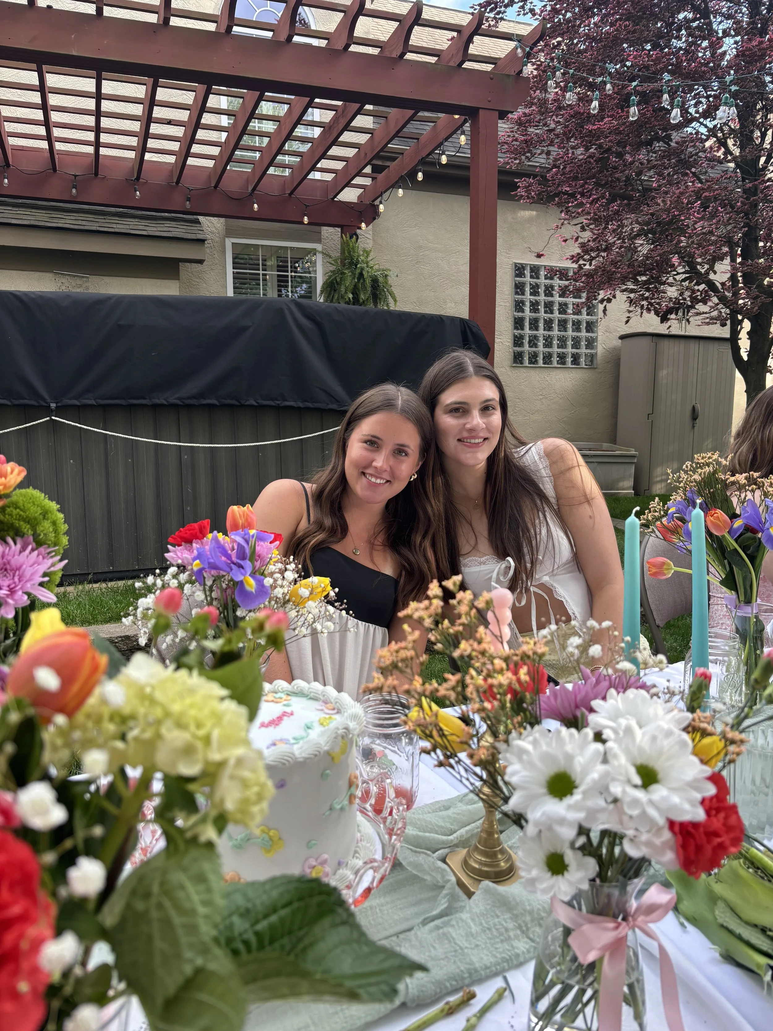 Two young women are sitting at a table decorated with colorful flowers, candles, and a birthday cake with a pink bow. They are smiling at the camera. The setting appears to be outdoors in a backyard with a wooden pergola, blooming tree, and exterior 