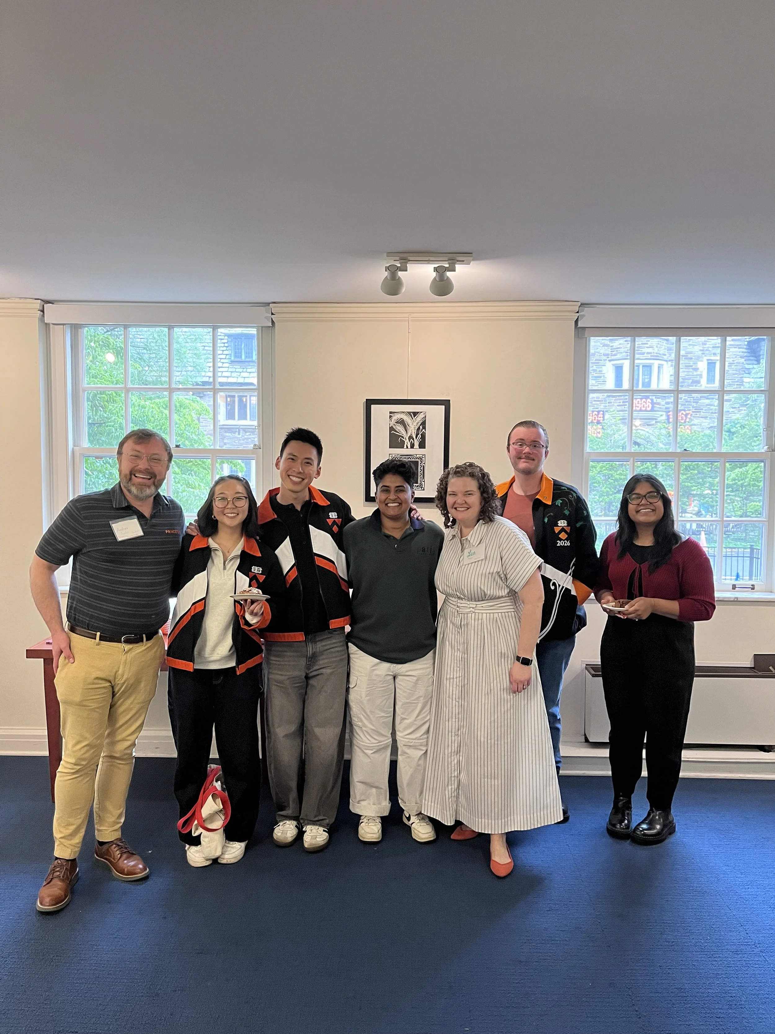 Group of seven people standing together indoors during Princeton Reunions, smiling and posing for a photo, with large windows in the background showing a building and trees outside.