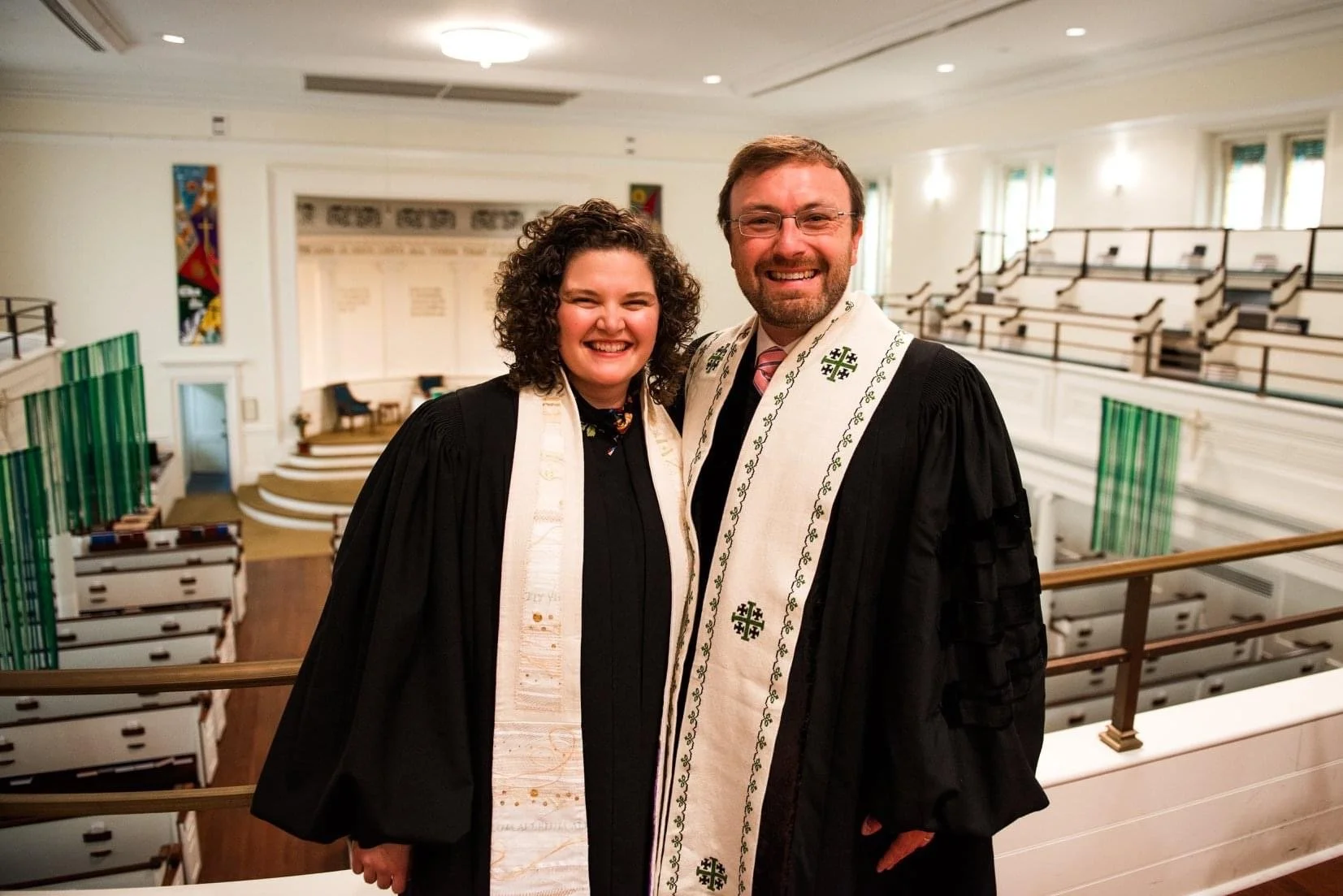 Two pastors in robes standing inside a church, smiling for a photo, with wooden pews and stained glass windows in the background.