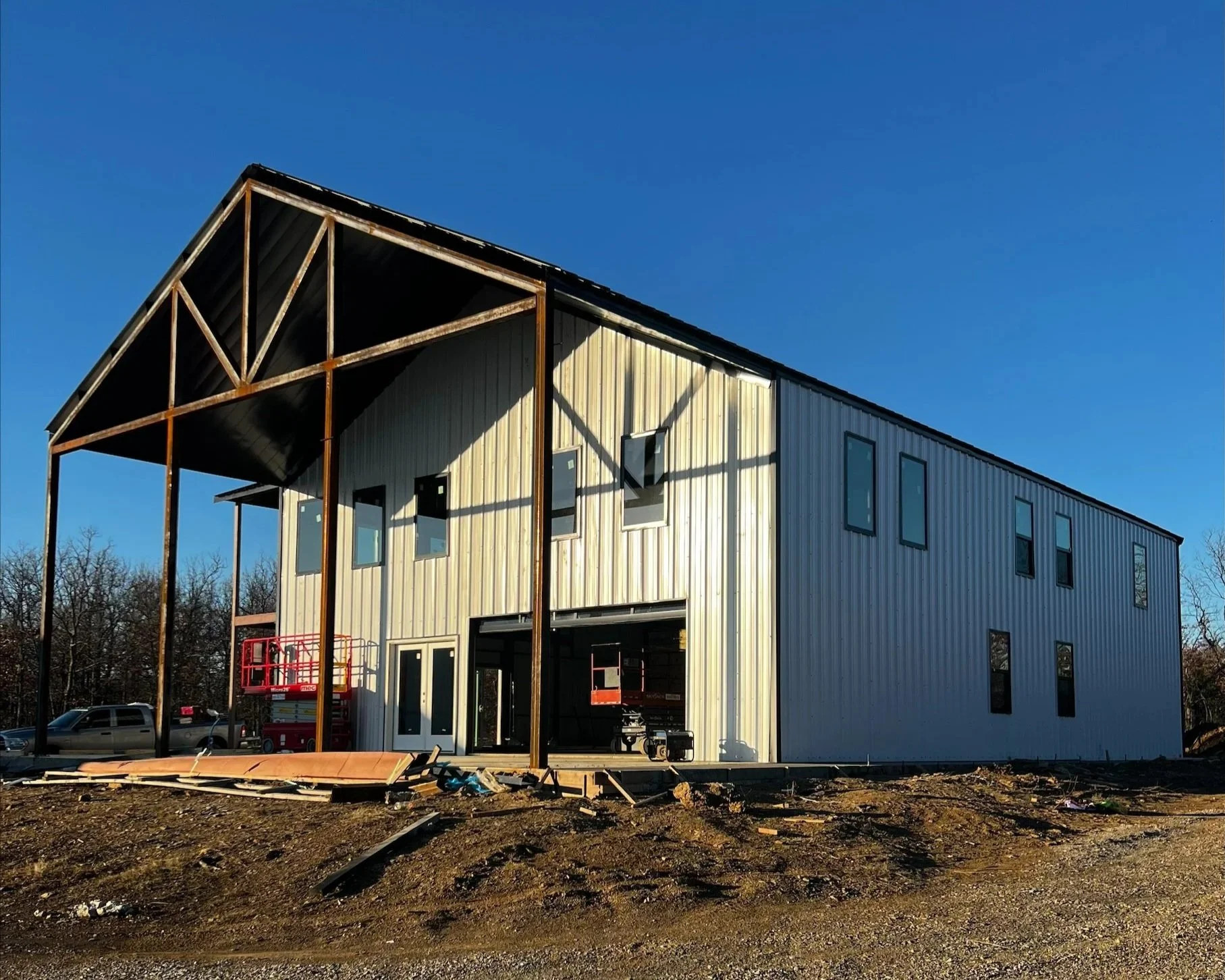 A building under construction with a metal exterior and large window openings, surrounded by construction debris and vehicles, with a blue sky overhead.