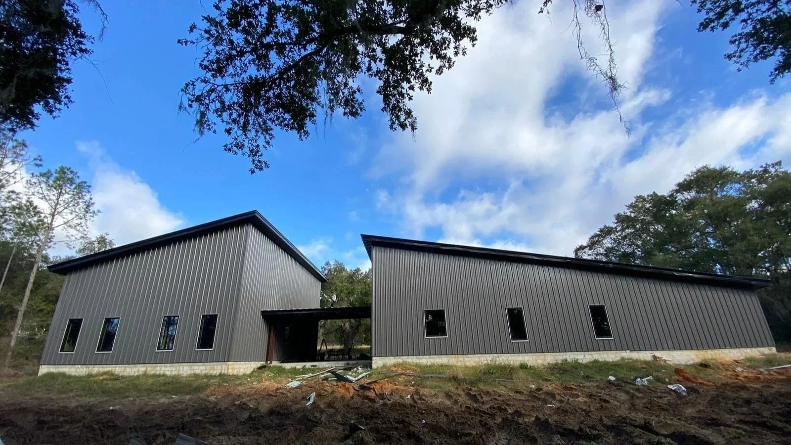 Two modern metal buildings with sloped roofs, situated in a wooded area under a partly cloudy sky.