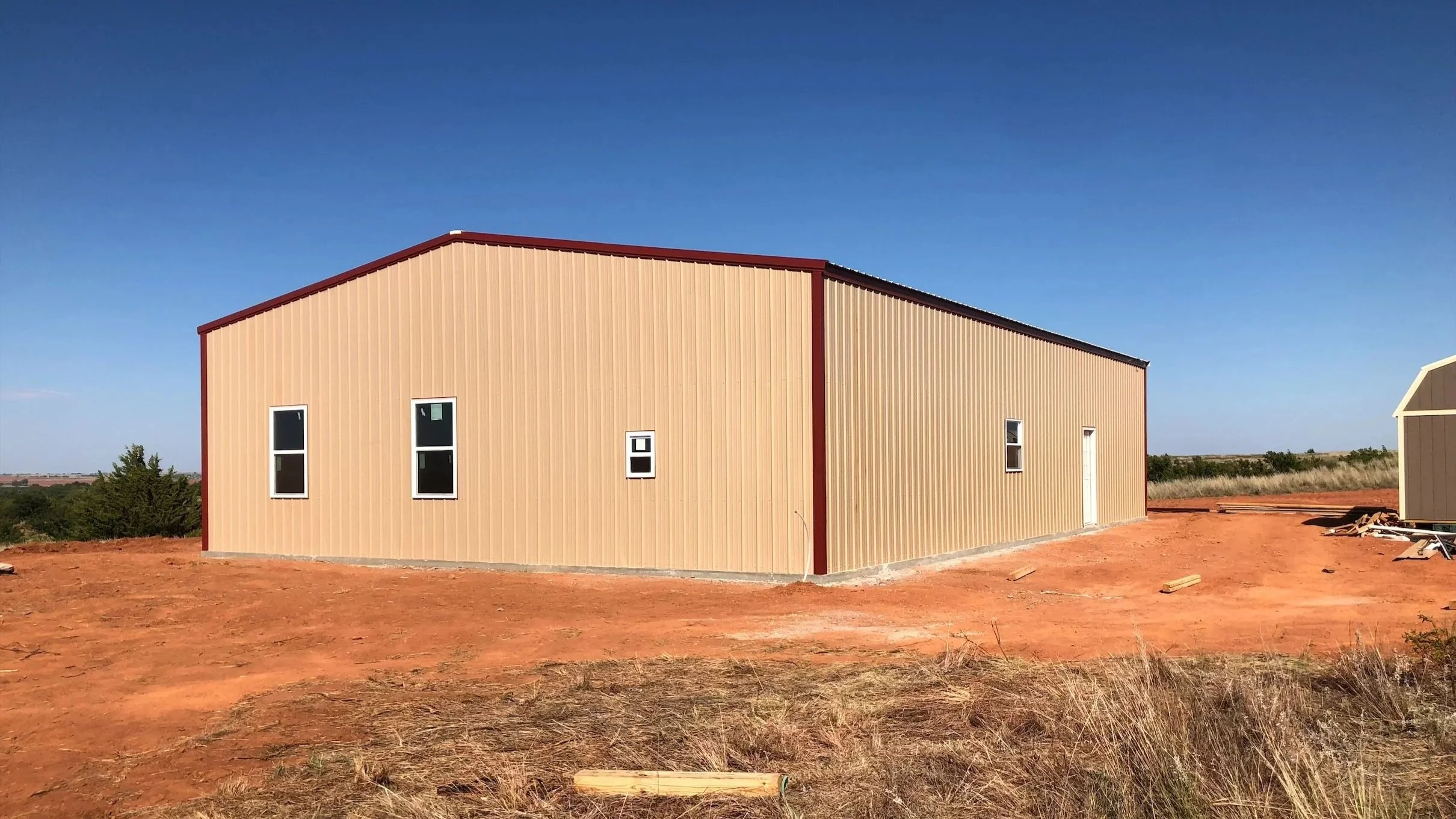 A beige metal building with four windows and a door, situated on a dirt lot under a clear blue sky, with some construction materials nearby.