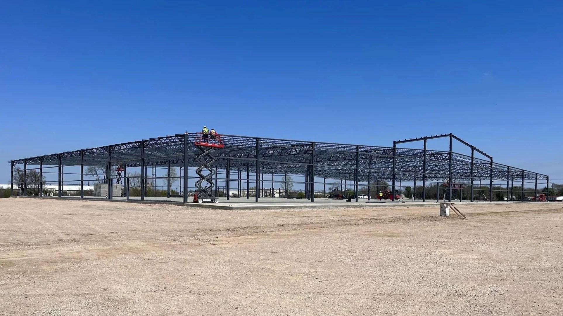 Construction workers on scissor lifts working on a large metal framework structure outdoors under a clear blue sky.