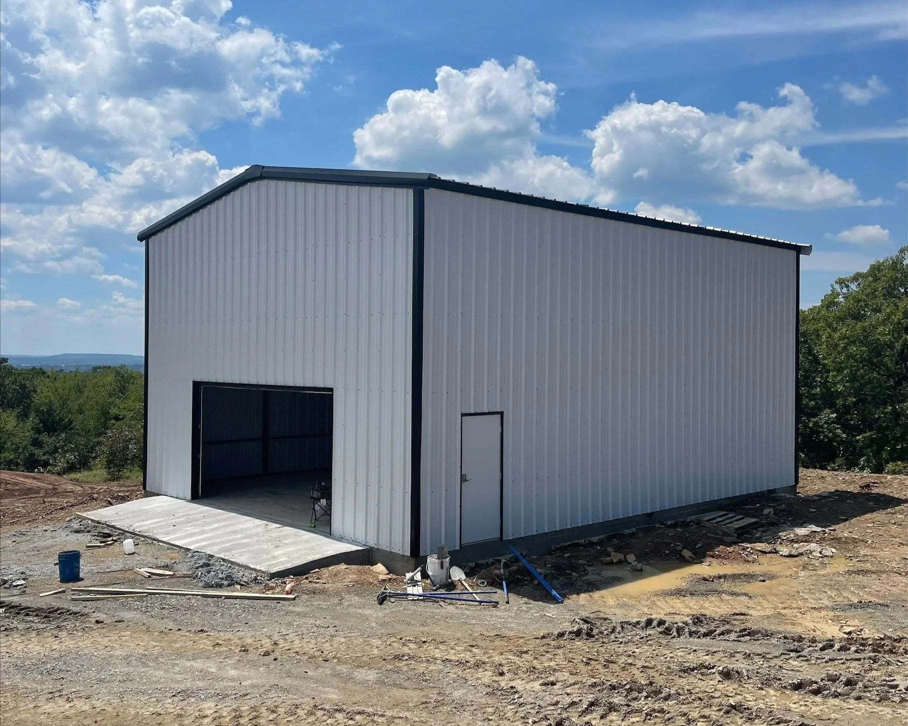 A metal building under construction with a white exterior, black trim, and open garage door, set against a partly cloudy sky with trees in the background.