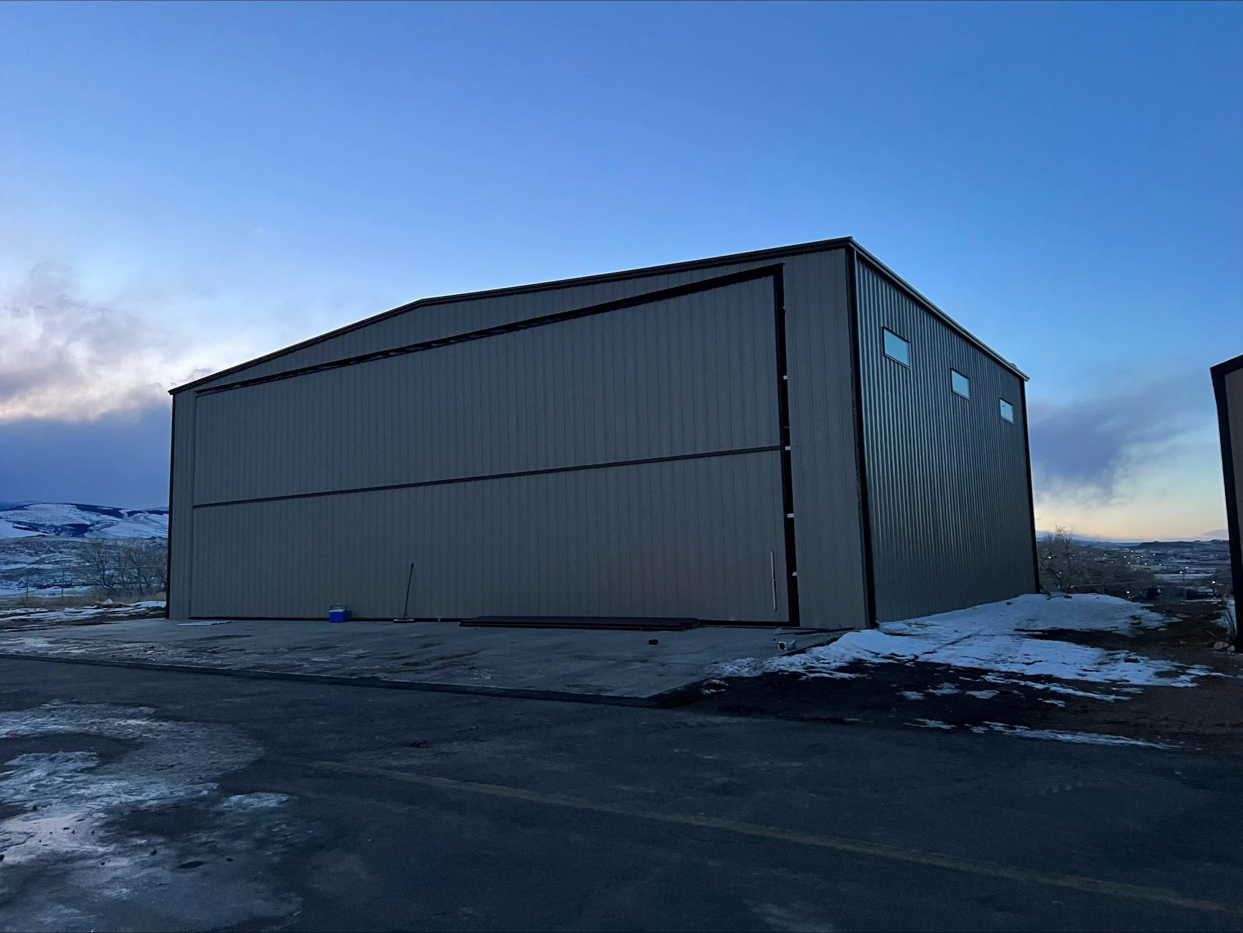 A large industrial building with a gray metal exterior, three small windows, and a sloped roof, situated in a snowy and cloudy landscape at dusk or dawn.